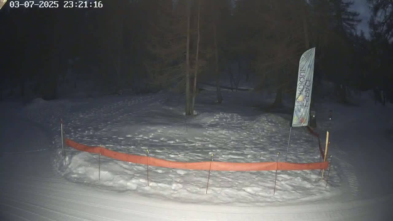 A snow-covered area at night, partially enclosed by an orange barrier, is dimly lit, with trees in the background.