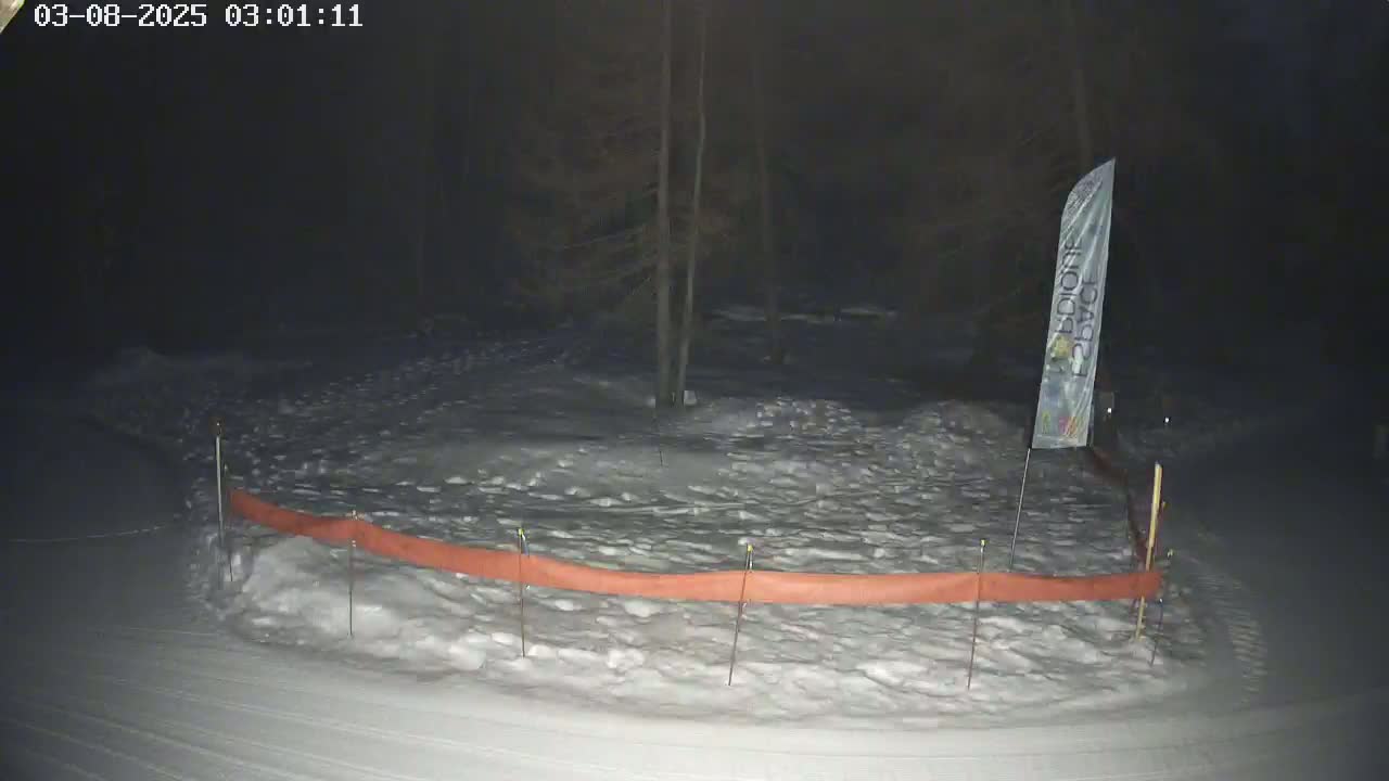A snow-covered area at night, illuminated by a faint light, is partially enclosed by an orange snow fence with a tall white banner standing nearby.