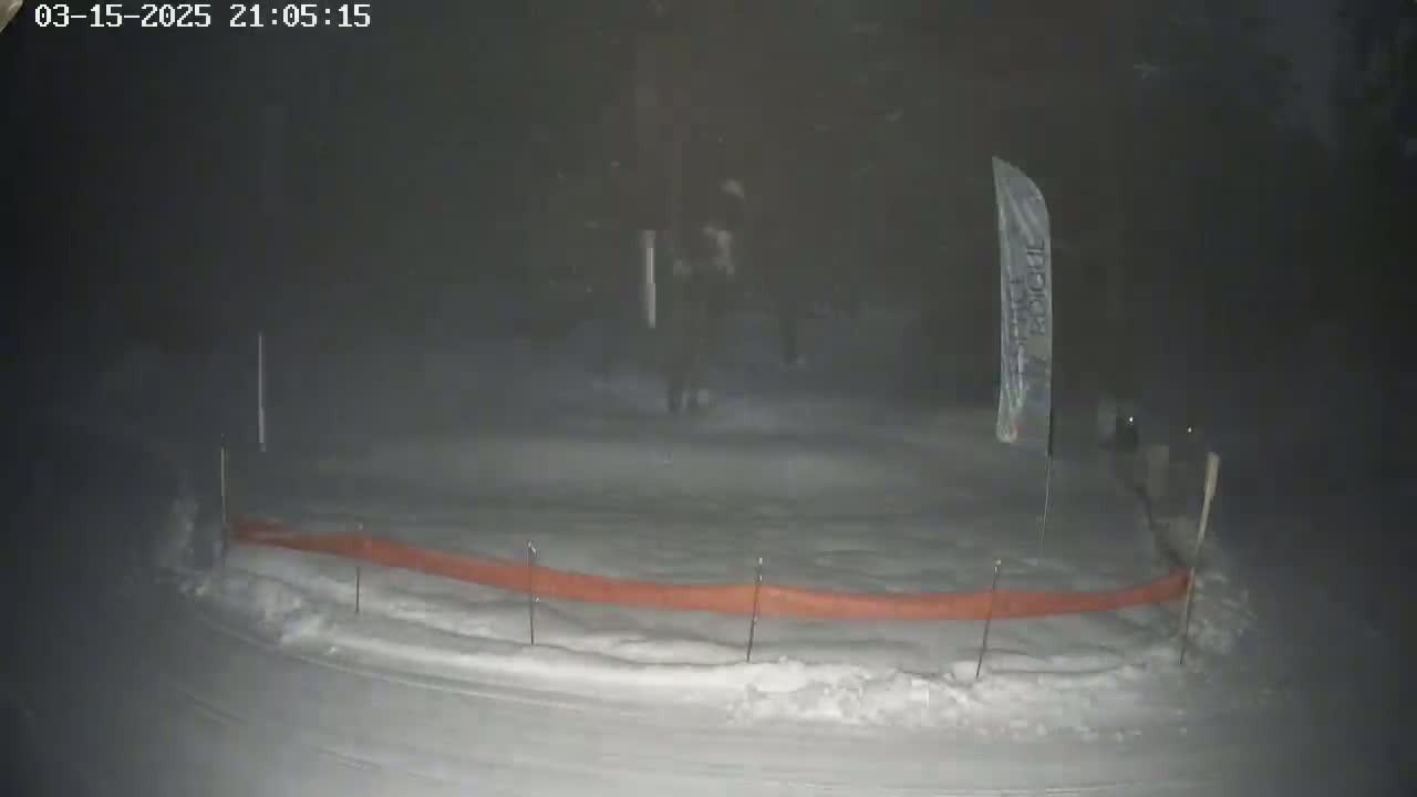 A snow-covered area at night, with an orange barrier and a lone flag visible in the falling snow.