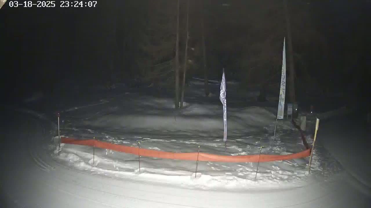 A snow-covered area at night, partially enclosed by an orange barrier, with several thin poles and flags visible.