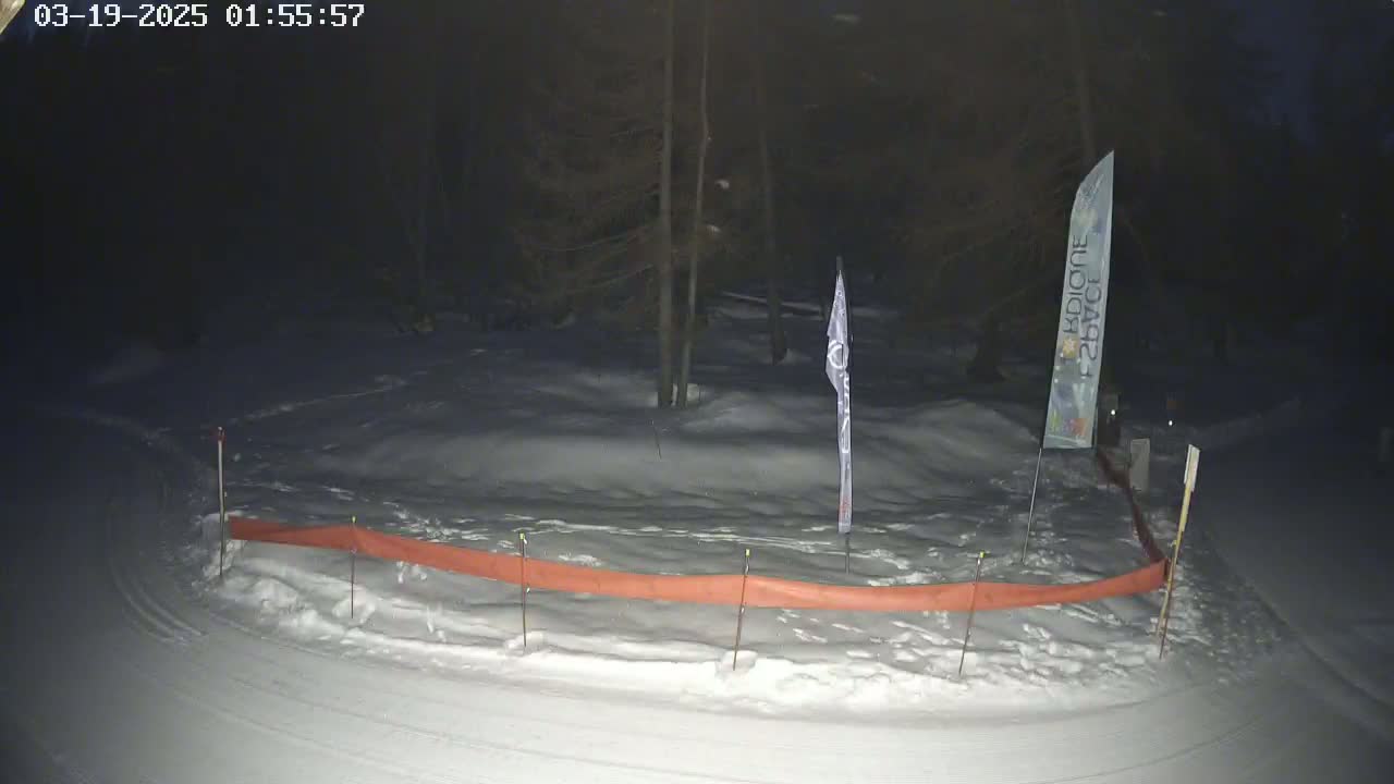 A snow-covered area at night, bordered by a low orange barrier, with a few trees and flags visible in the background.