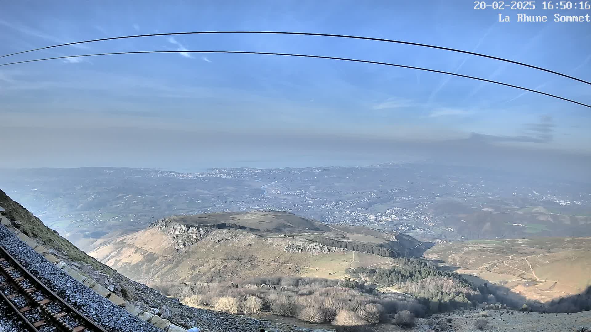 A mountaintop view shows a vast expanse of land, a distant body of water, a railway track beside a rocky slope, and a clear, sunny sky.