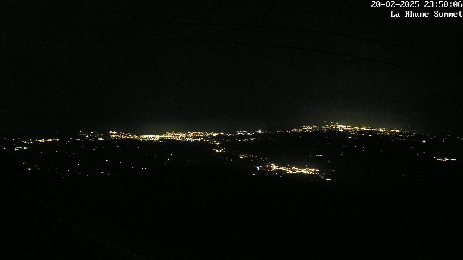 A nighttime long shot shows a sprawling city illuminated by countless lights, nestled in a dark, hilly landscape under a clear, black sky.