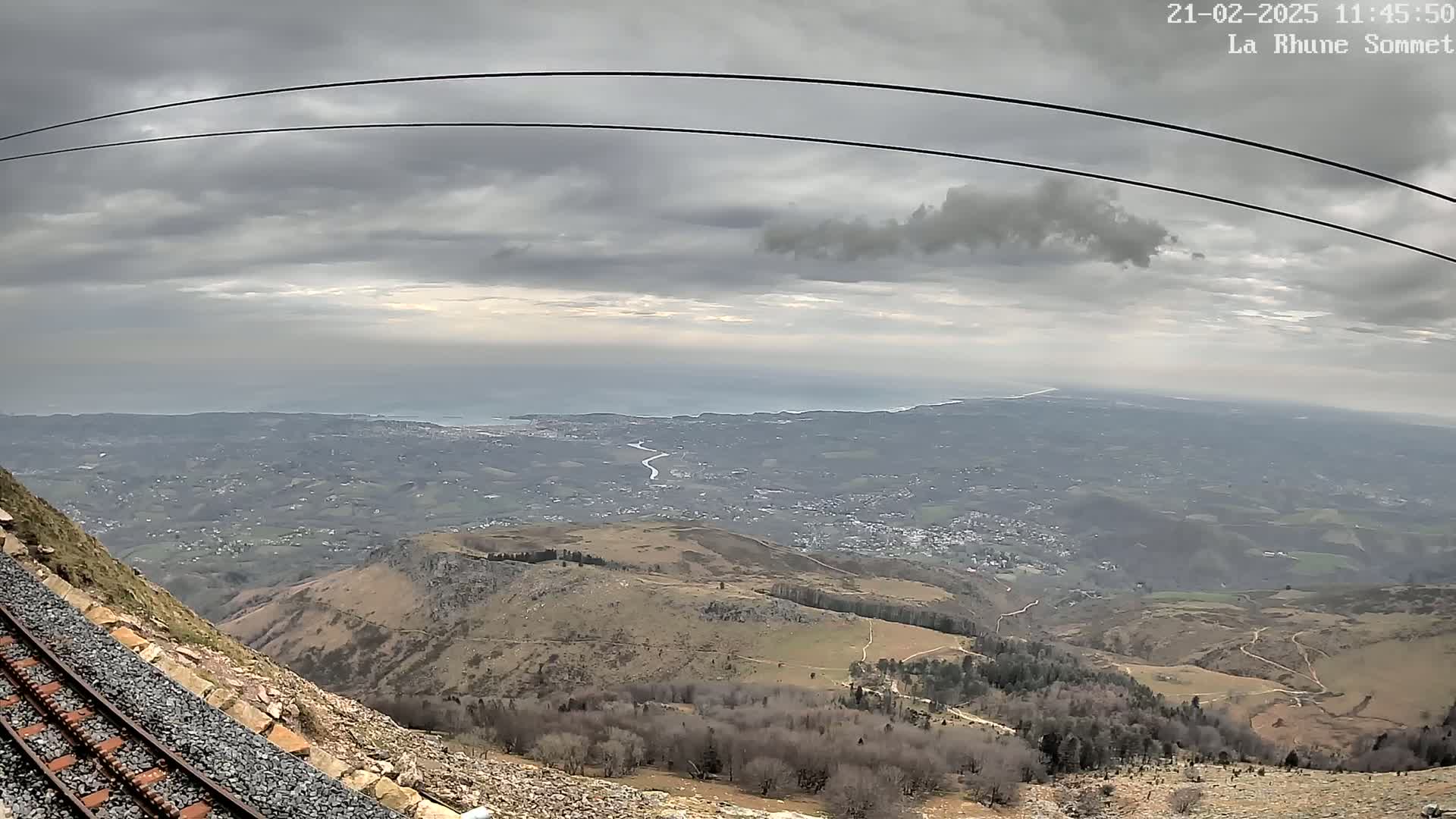 A mountaintop view shows a vast expanse of land extending to a distant ocean under a cloudy sky, with a section of railway tracks in the foreground.