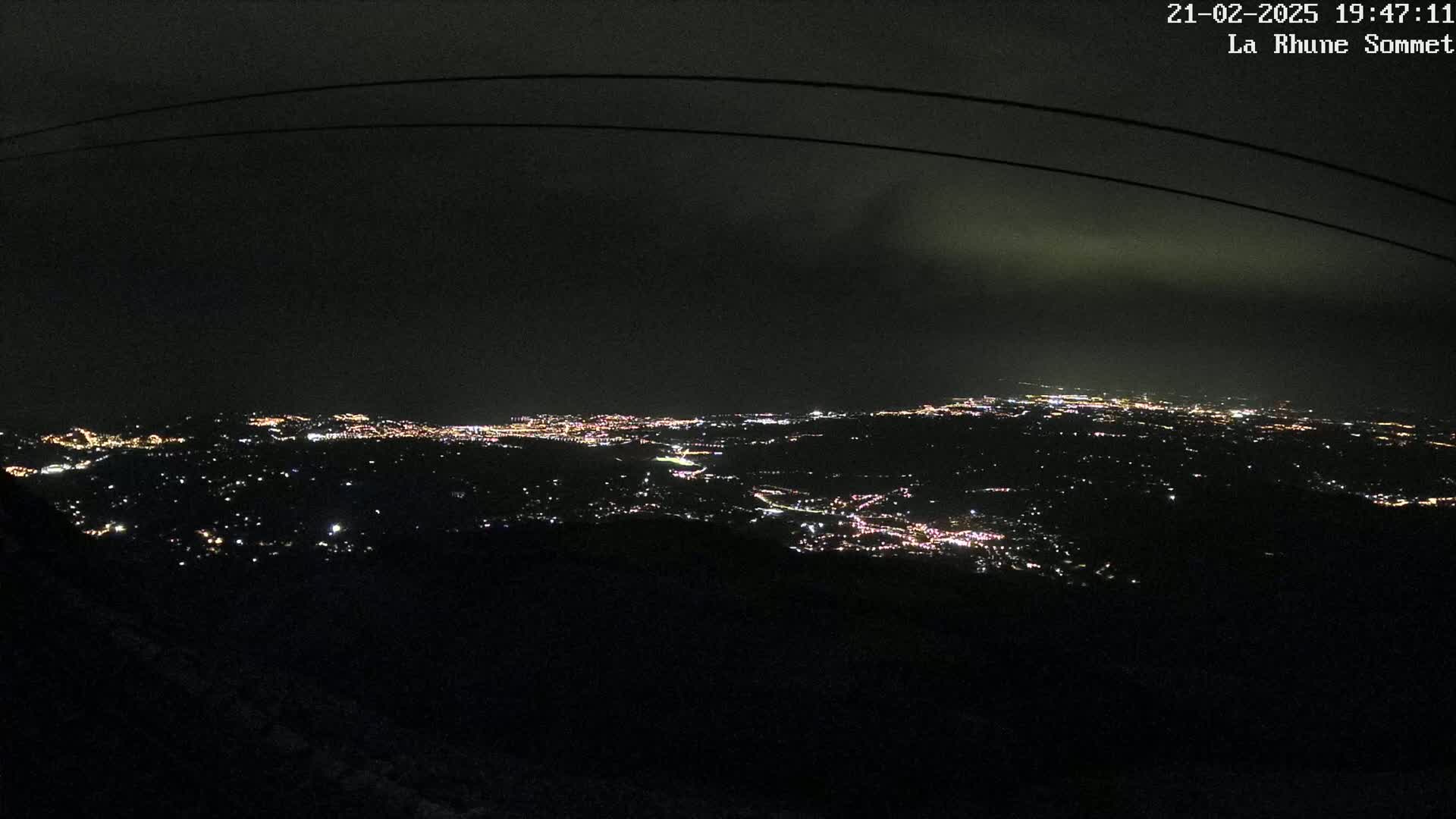 A nighttime view of a sprawling city illuminated by countless lights, seen from a high vantage point under a dark, cloudy sky.