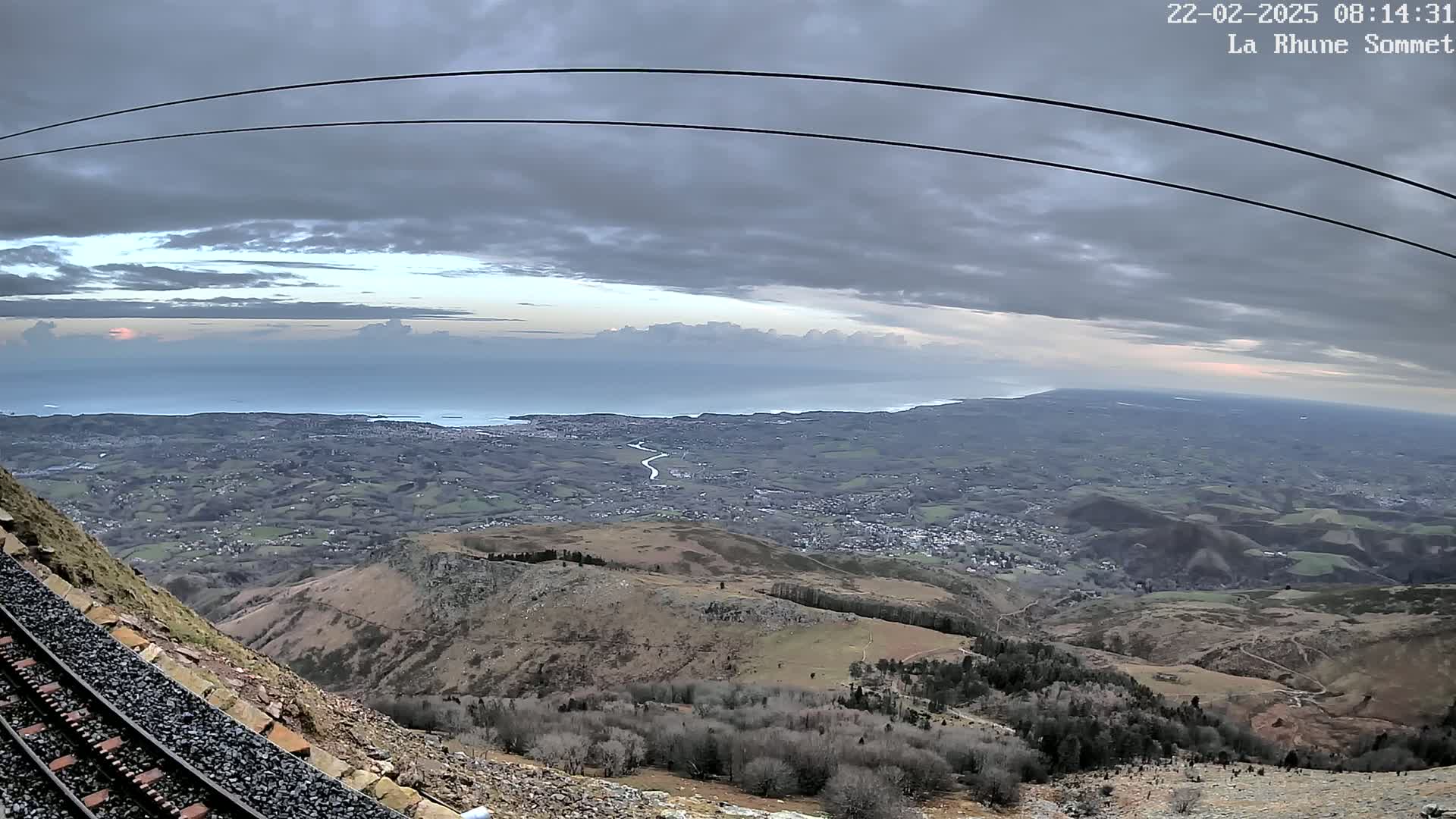 A mountaintop view reveals a railway track beside a rocky slope overlooking a sprawling coastal landscape under an overcast sky.