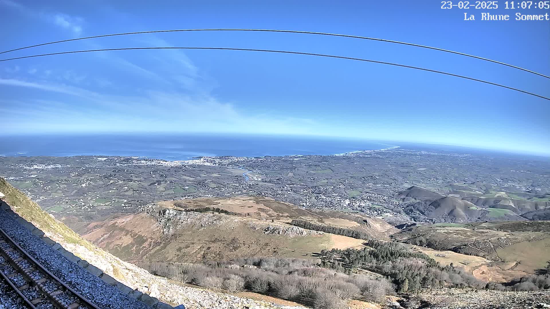 A mountaintop view on a clear, sunny day shows a vast expanse of land extending to a distant ocean, with a railway track visible in the foreground.