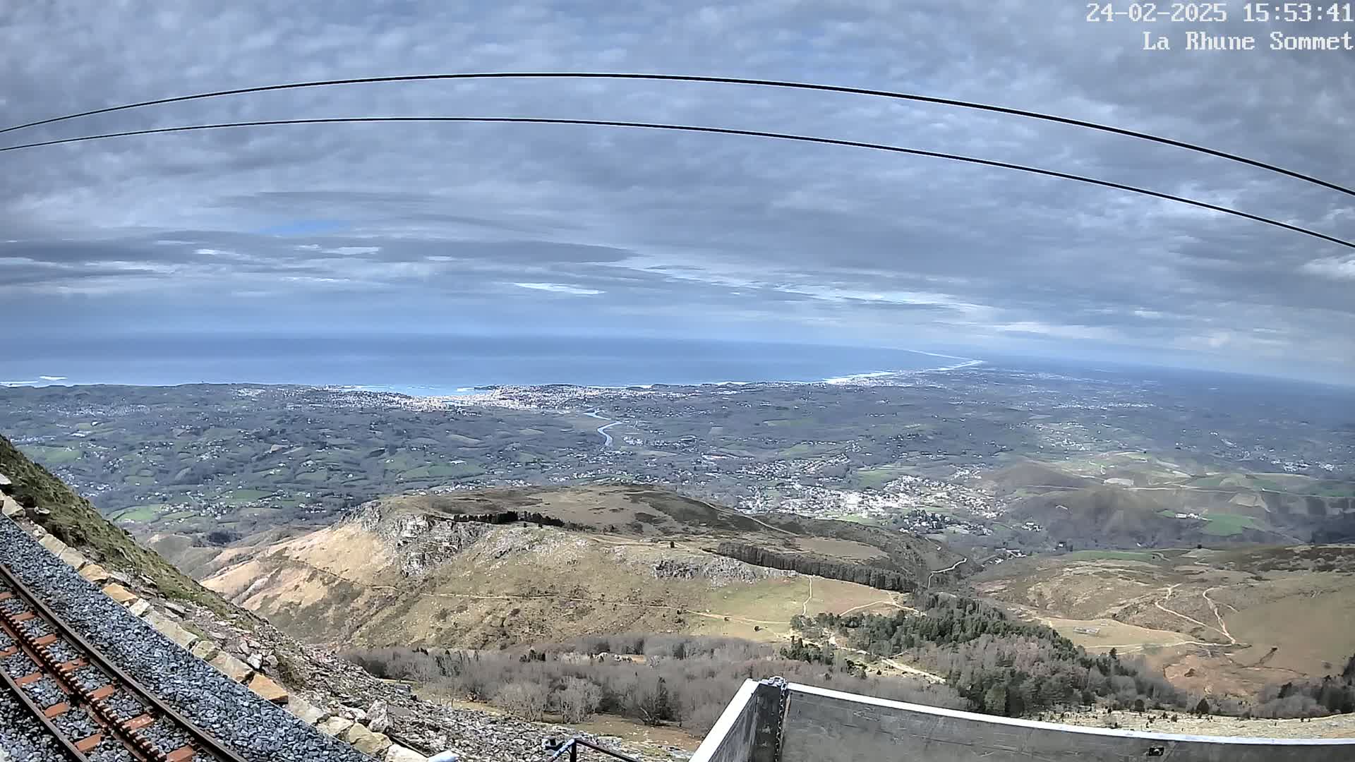 A mountaintop railway track overlooks a sprawling coastal landscape under a cloudy sky.