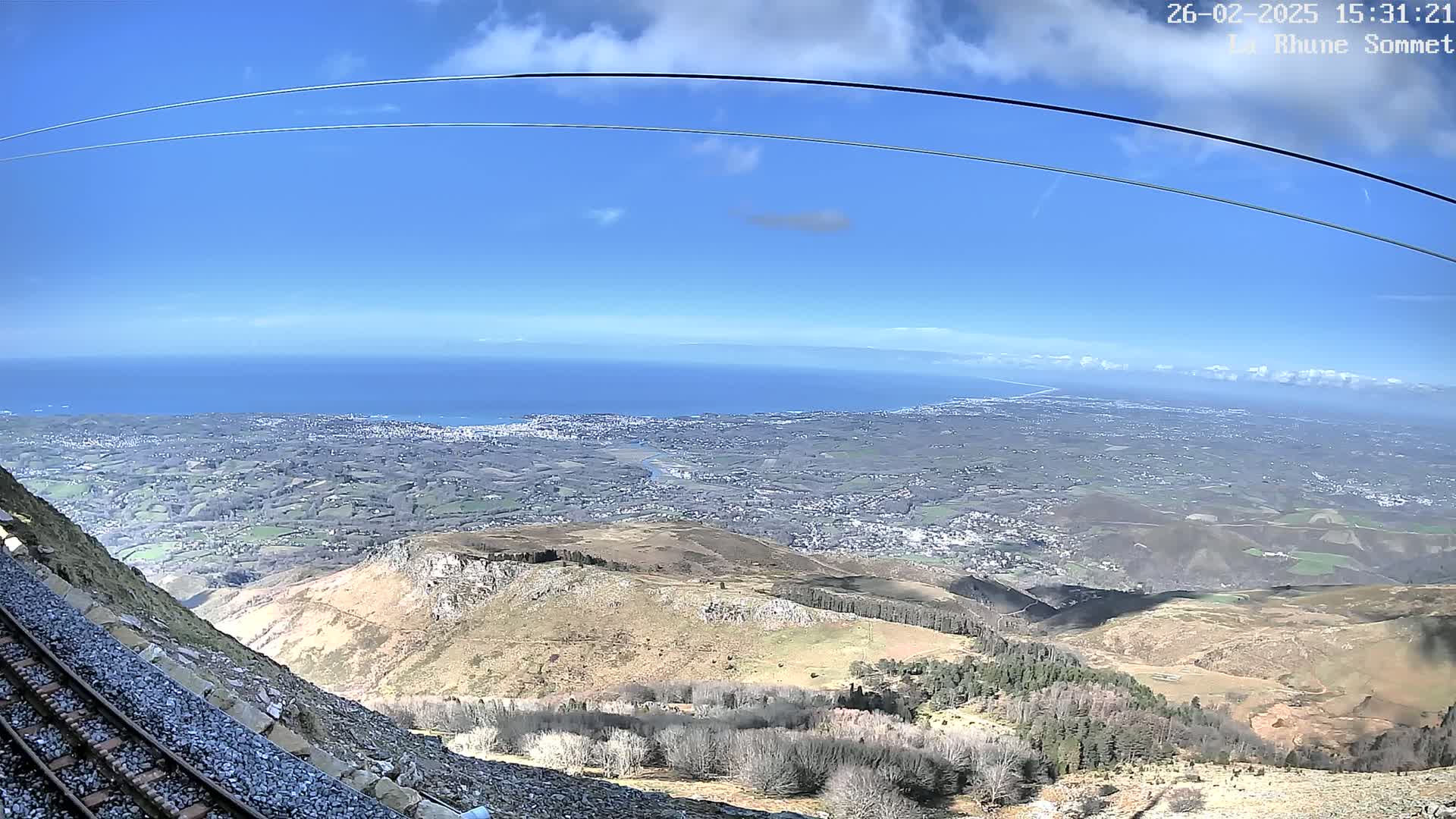 A mountaintop view on a clear, sunny day shows a vast expanse of land, a town in the distance, and a body of water beyond, all visible from a railway track running alongside a steep hillside with sparse vegetation.