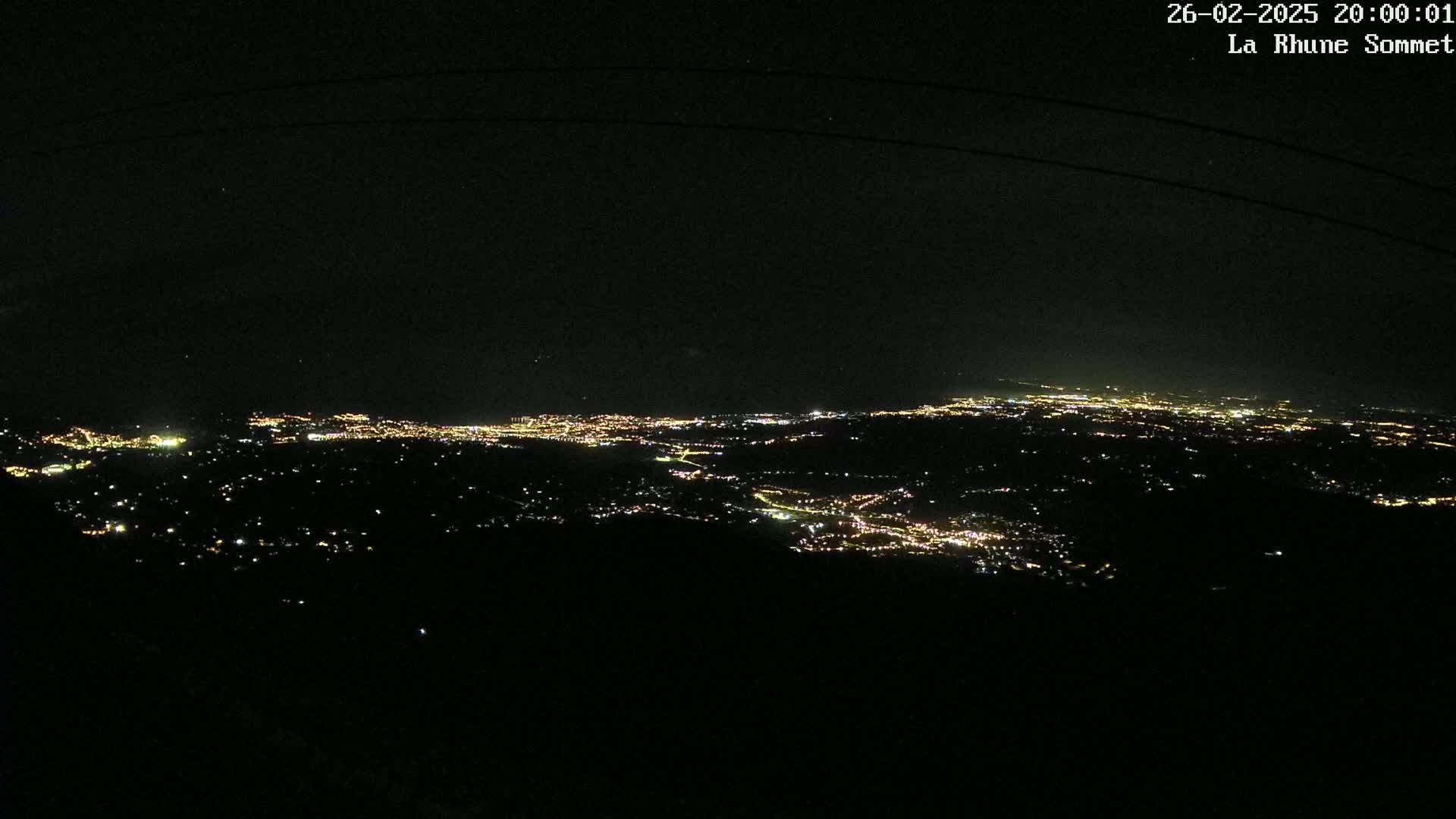 A nighttime, high-angle view shows a sprawling city illuminated by numerous lights, extending across a dark landscape under a clear sky.
