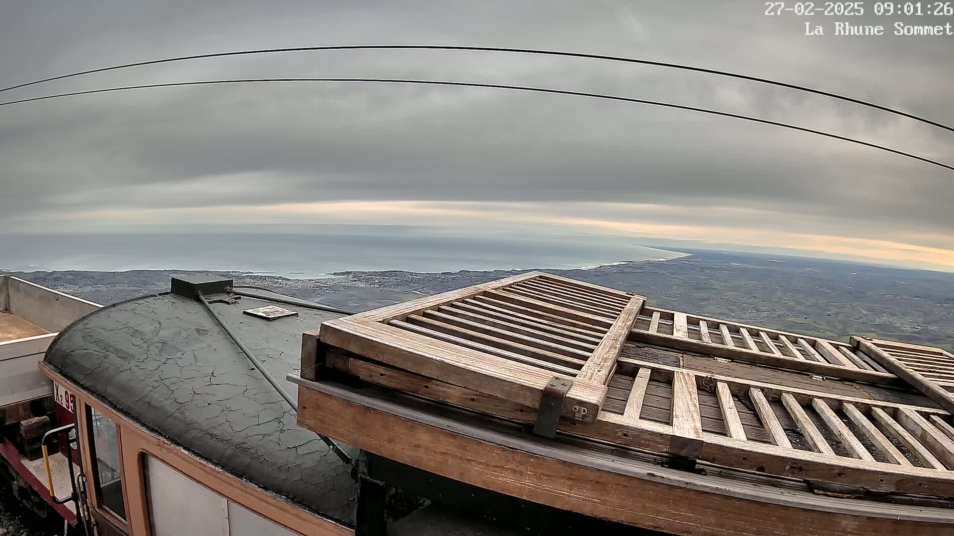 A weathered train car with wooden slats on its roof sits atop a hill overlooking a calm ocean under a cloudy sky.