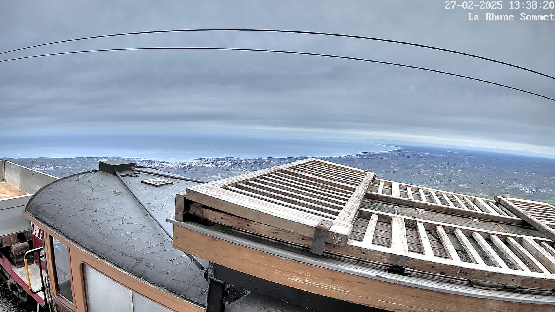 A train car with weathered wooden slats on its roof is parked overlooking a coastal town and ocean under a cloudy sky.