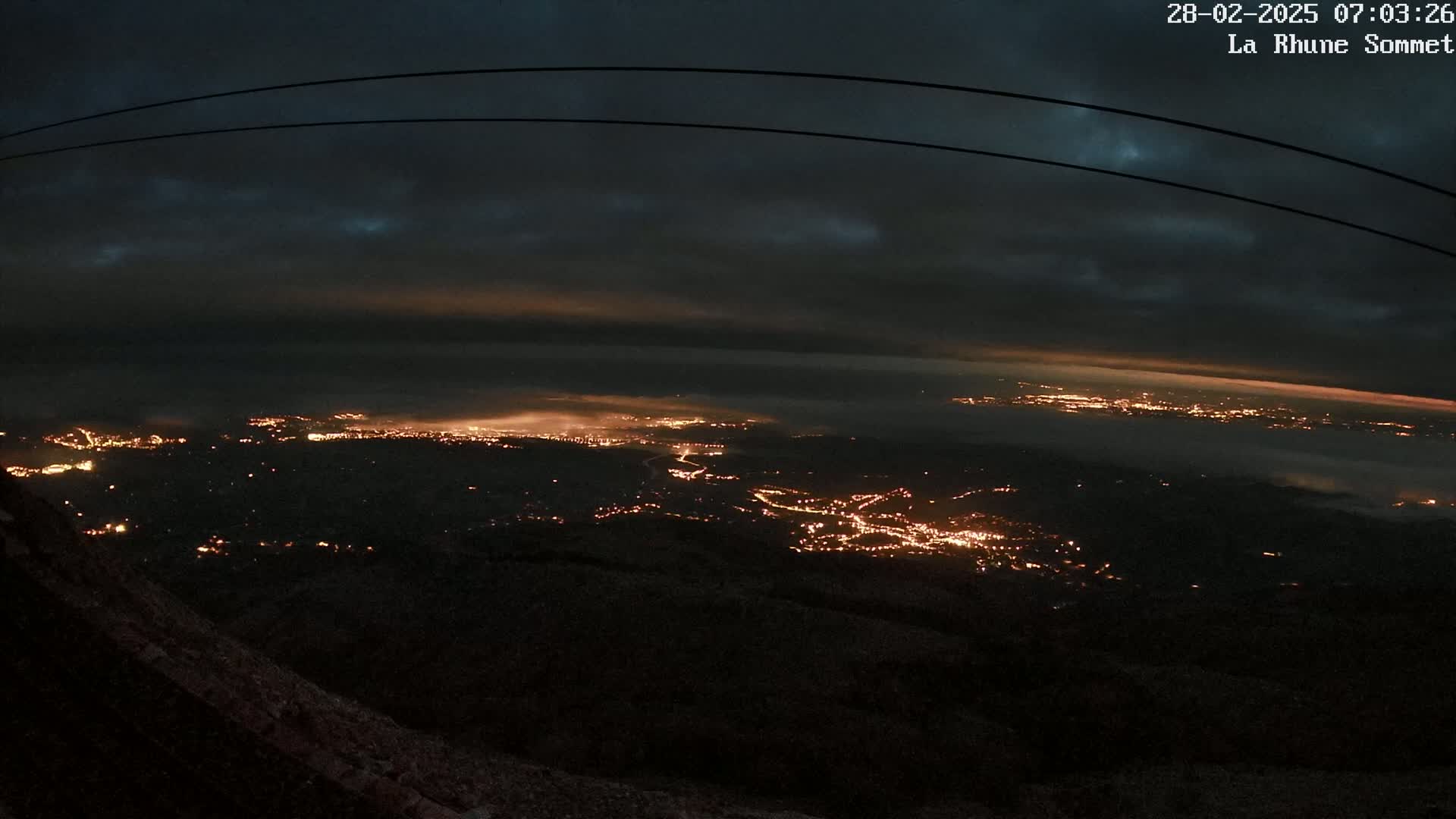 A nighttime view from a high vantage point shows a sprawling city illuminated below, partially obscured by a low layer of fog or clouds under a dark, cloudy sky.