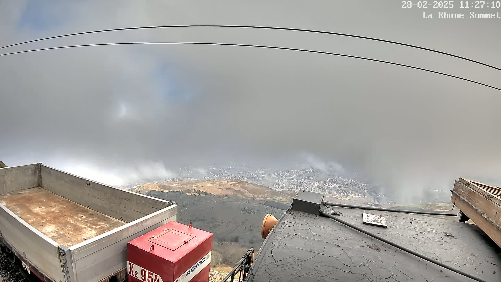 A view from a high vantage point shows a foggy landscape with a town below, partially obscured by clouds, and two pieces of industrial equipment, one a flatbed truck and the other a dark-colored structure, in the foreground.
