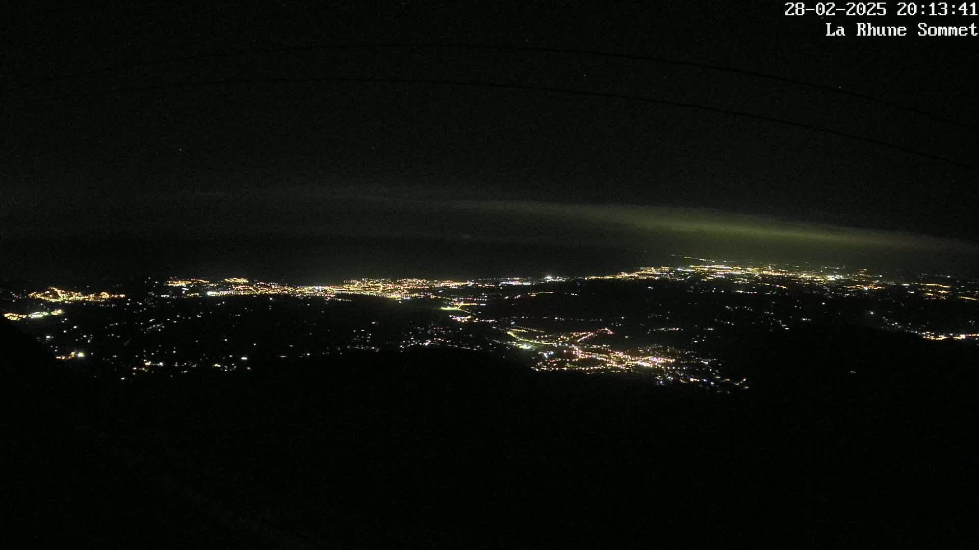 A nighttime, high-angle view shows a sprawling cityscape illuminated by numerous lights under a dark, slightly hazy sky.