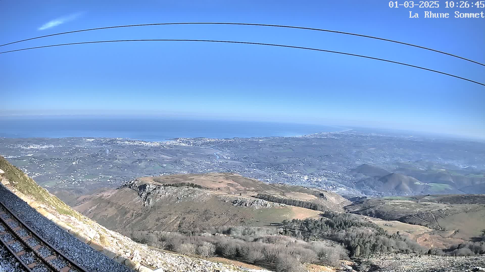 A mountaintop view on a clear day shows a vast expanse of land stretching to a distant ocean, with train tracks and barren hills in the foreground.