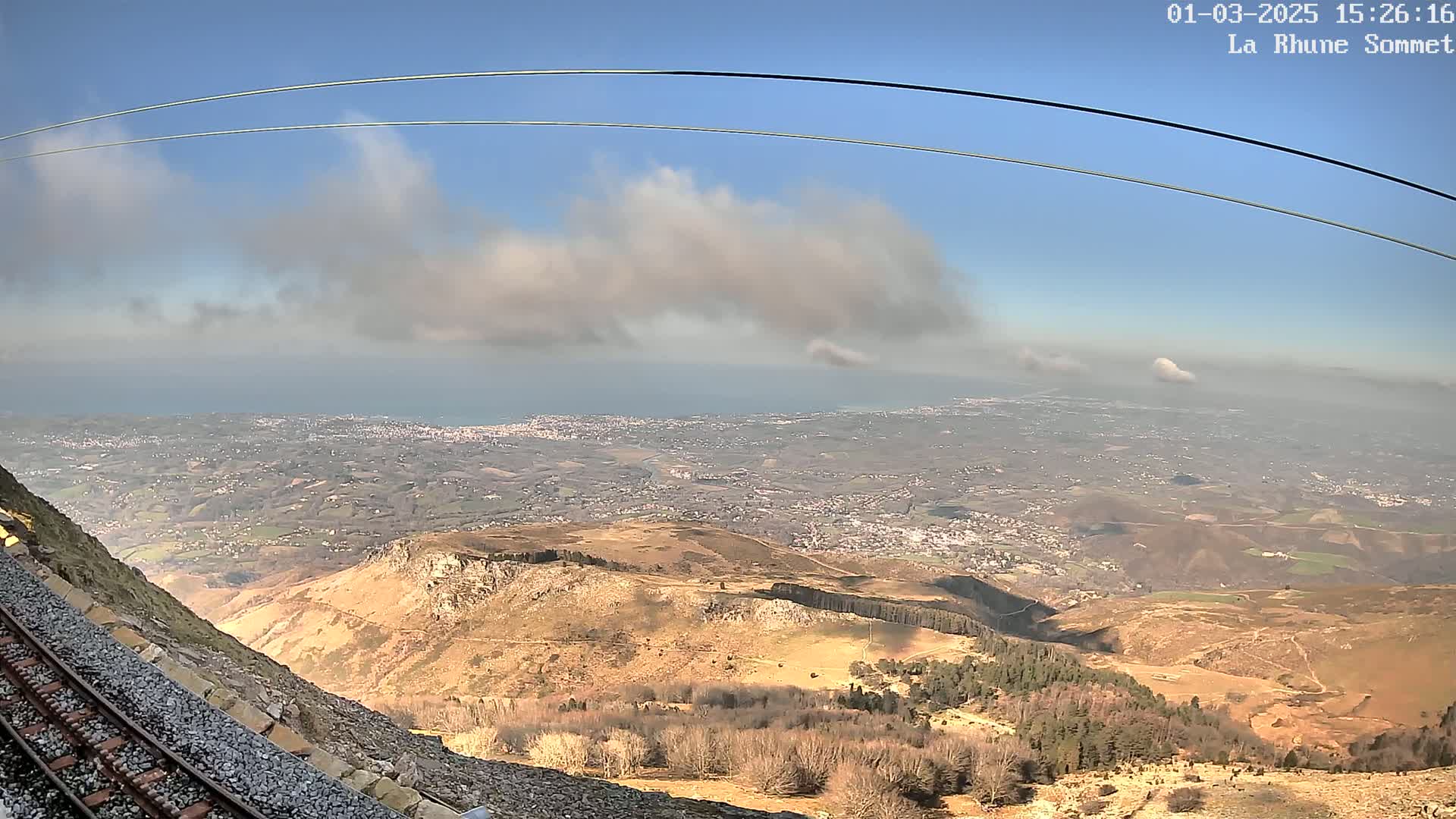 A mountaintop view on a partly cloudy day shows a vast expanse of land, including a distant coastline and a town below, with a railway track visible in the foreground.