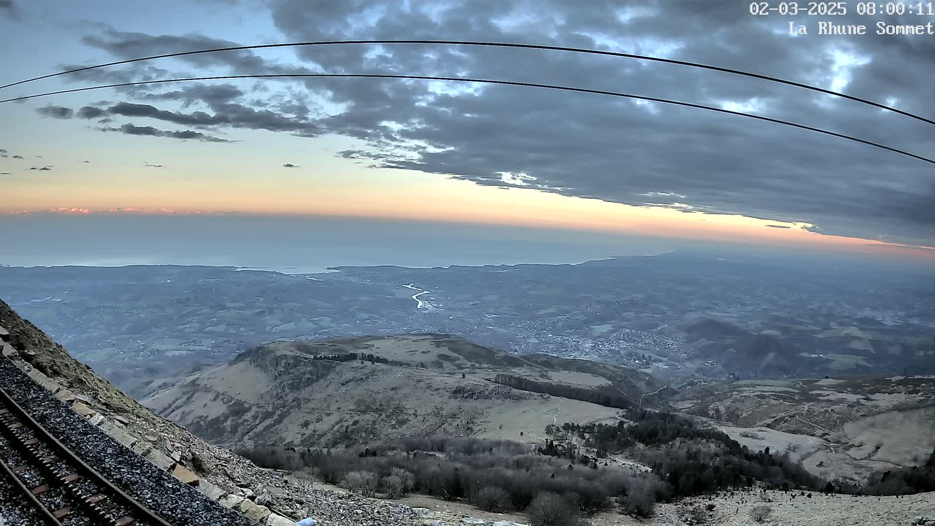 A mountaintop view at sunrise shows a vast expanse of land extending to a calm ocean under a partly cloudy sky, with a railway track running alongside a rocky slope in the foreground.