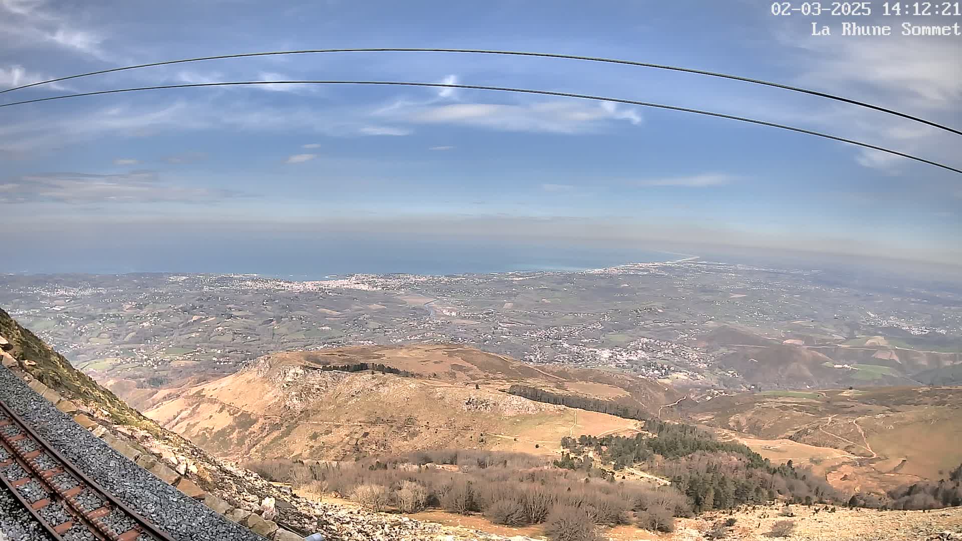A mountaintop view reveals a vast expanse of land stretching to a distant ocean under a mostly clear, bright sky, with train tracks visible in the foreground.