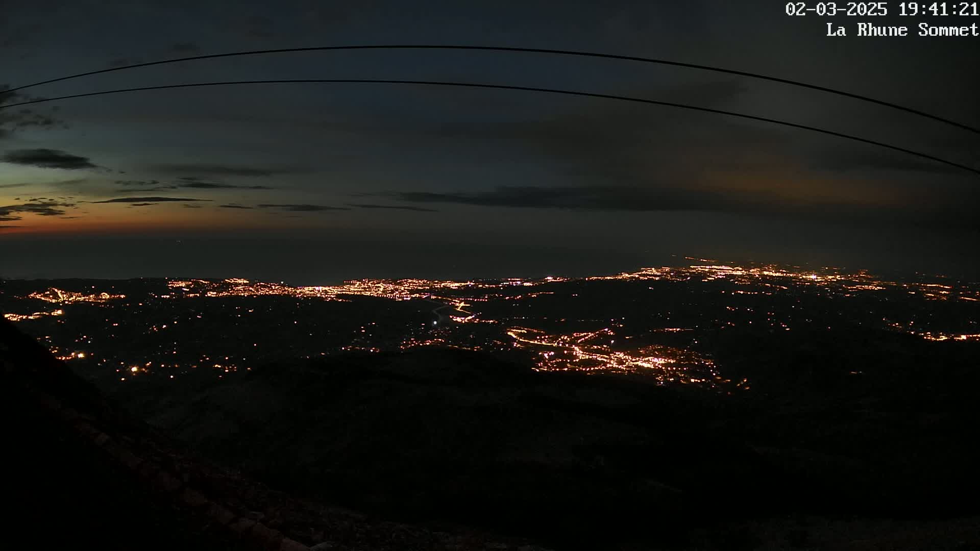 A nighttime, high-angle view shows a sprawling city illuminated with numerous lights, extending towards a dark ocean under a mostly clear, dusky sky.