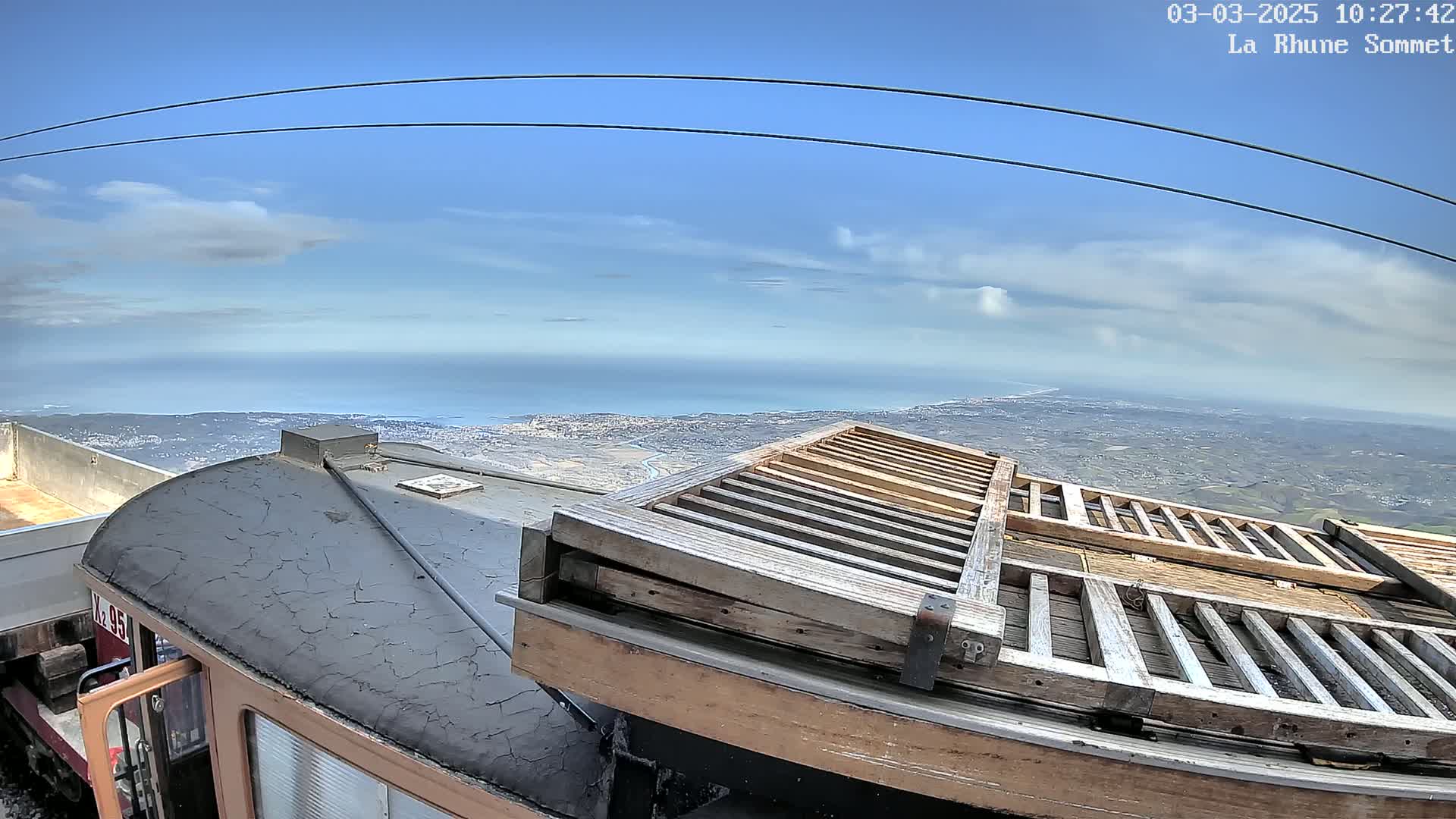 A train car's roof and a wooden structure are visible, overlooking a coastal town and ocean under a mostly clear sky.