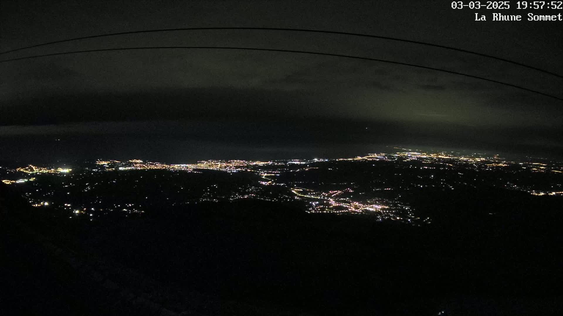 A nighttime, high-angle view of a sprawling city illuminated by numerous lights beneath a dark, cloudy sky.