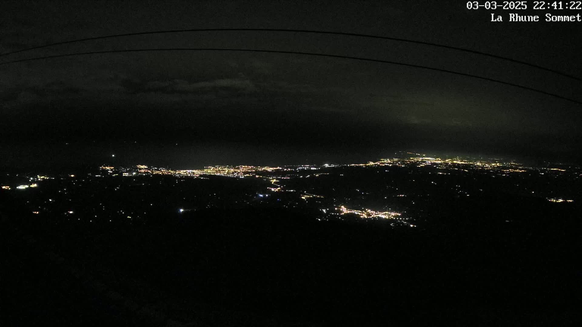 A nighttime, high-angle view shows a sprawling city's lights spread across a dark landscape under a cloudy sky.