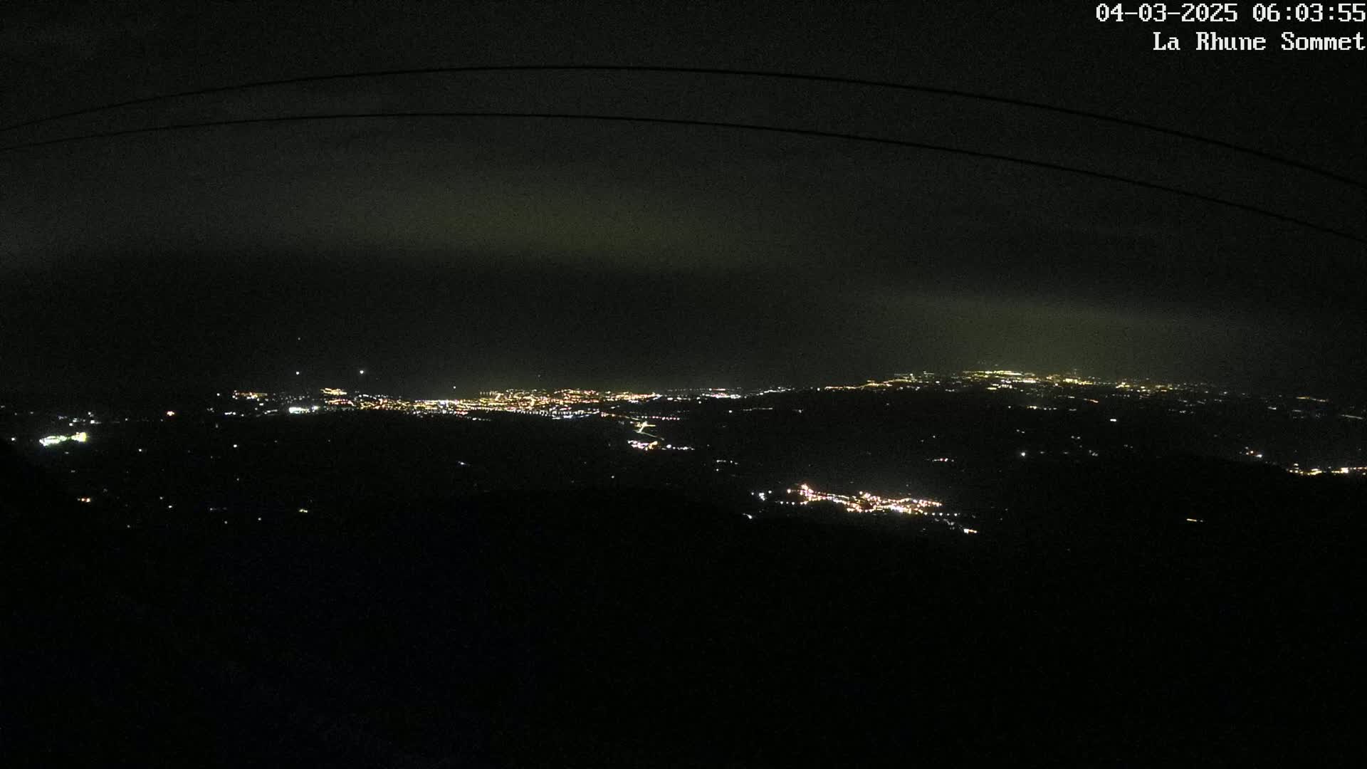 A nighttime view of a distant city's lights spread across a dark landscape under a cloudy sky.