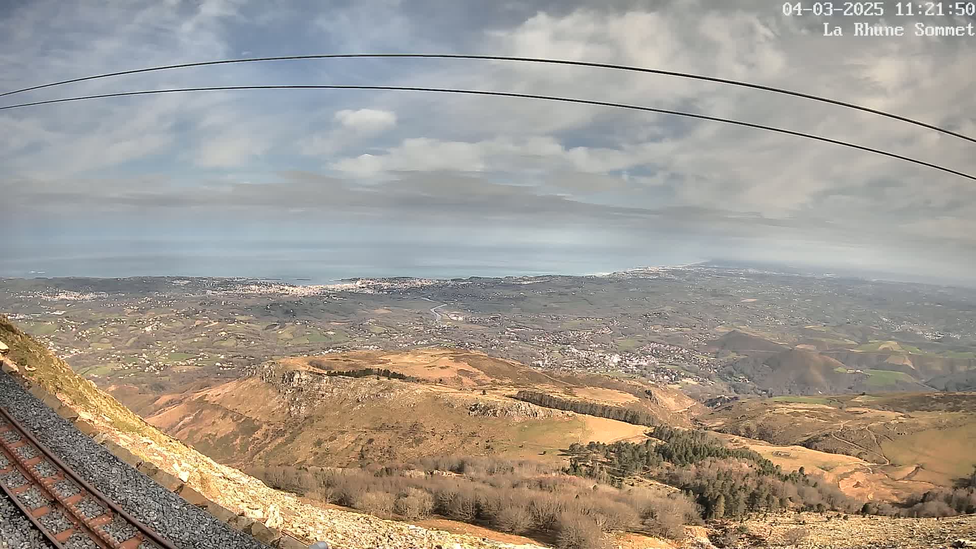 A mountaintop view shows a coastal town and ocean under a partly cloudy sky, with a railway track and cable lines in the foreground.