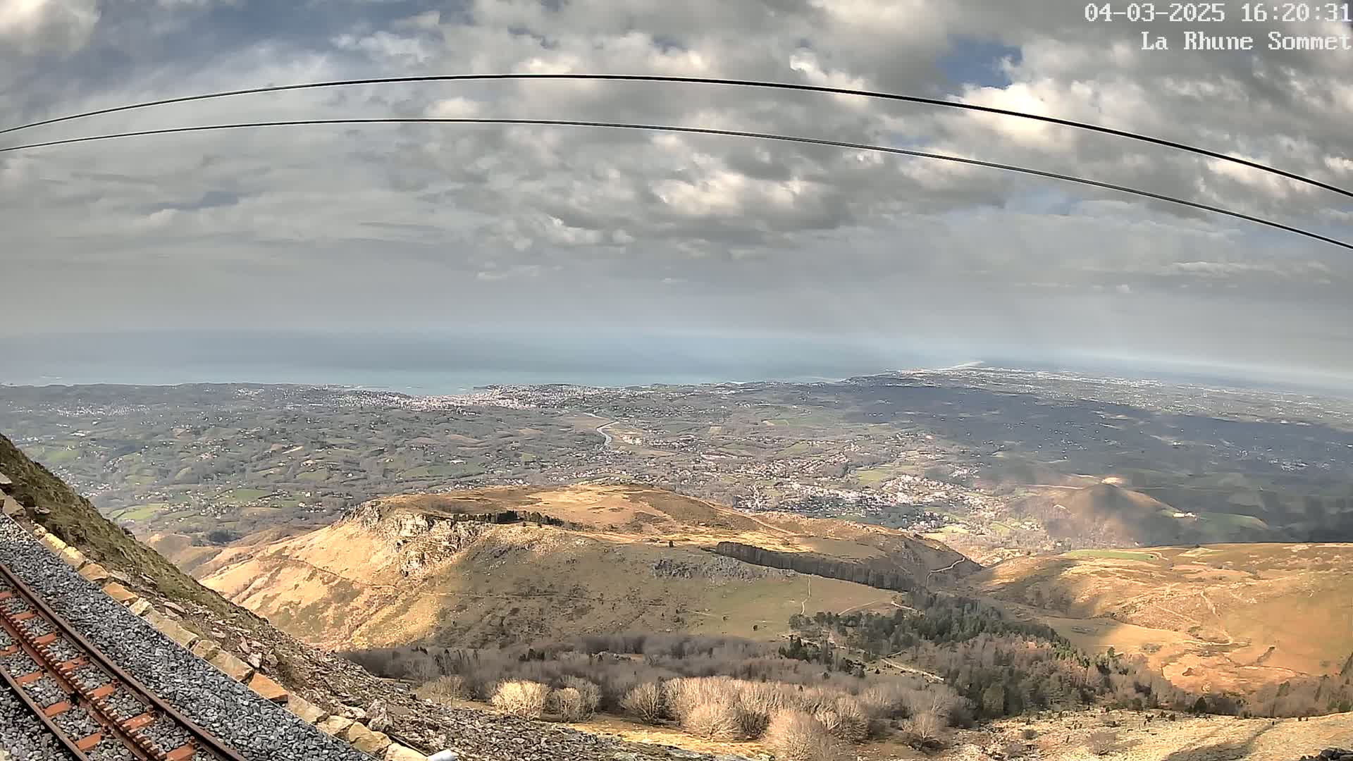 A mountain railway track alongside a rocky hillside overlooks a vast expanse of land, a distant coastline, and a partly cloudy sky.