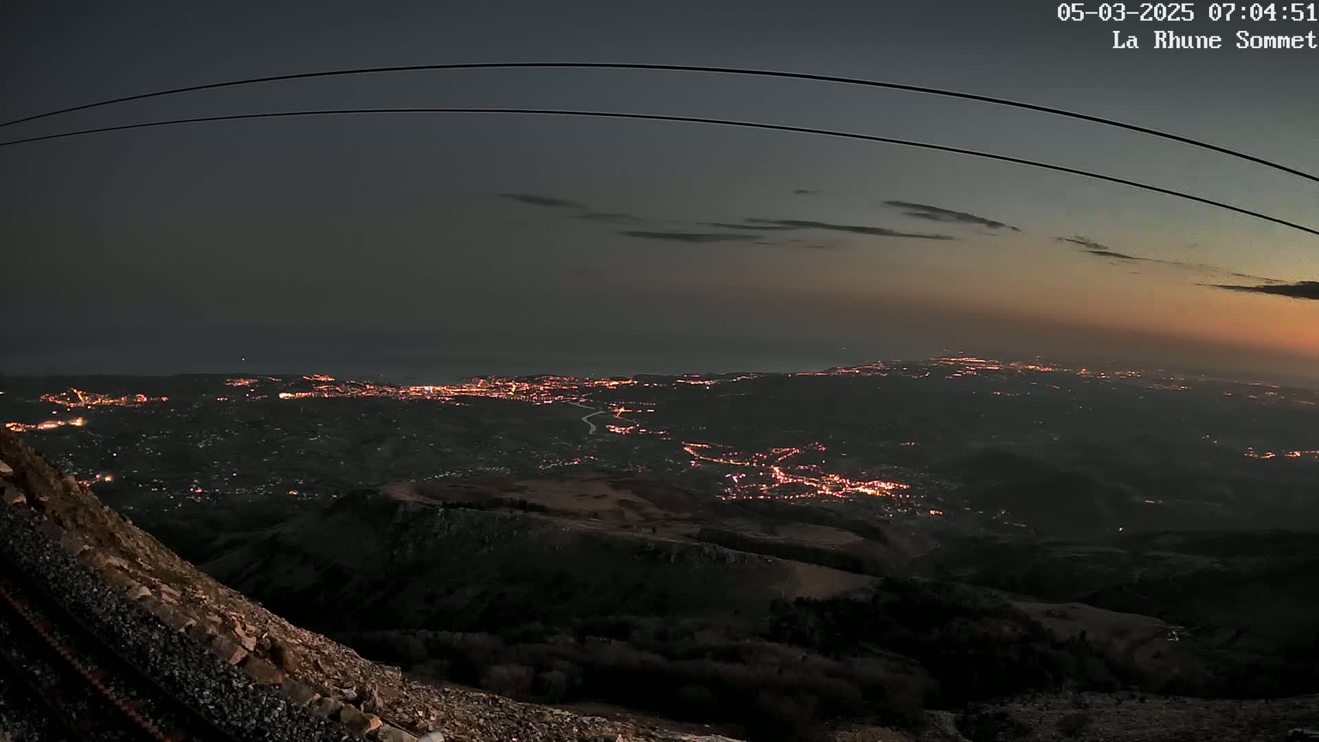 A nighttime view from a mountaintop shows a sprawling city illuminated below, with a dark ocean visible in the distance under a dusky sky.