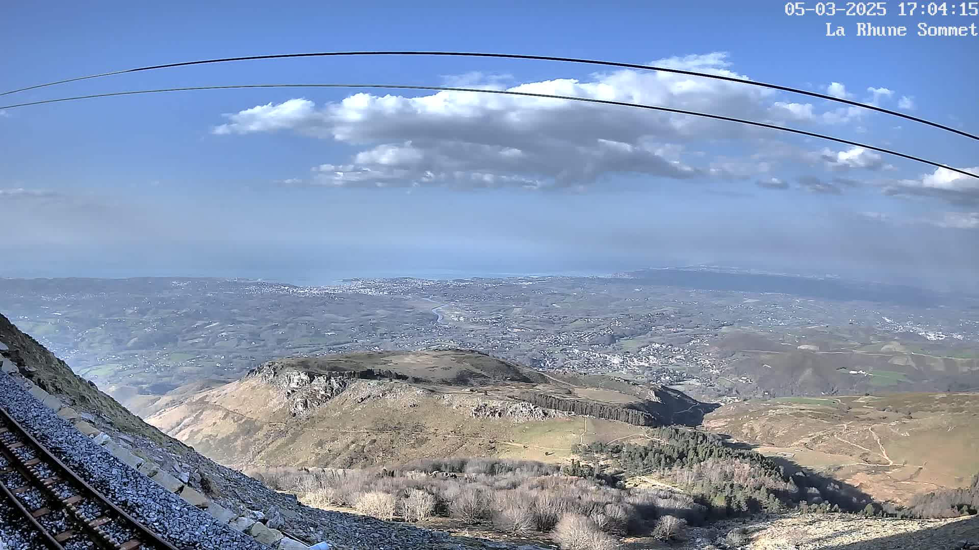 A mountaintop view shows a sprawling landscape, a distant ocean, and a railway track alongside a steep, rocky incline under a partly cloudy sky.