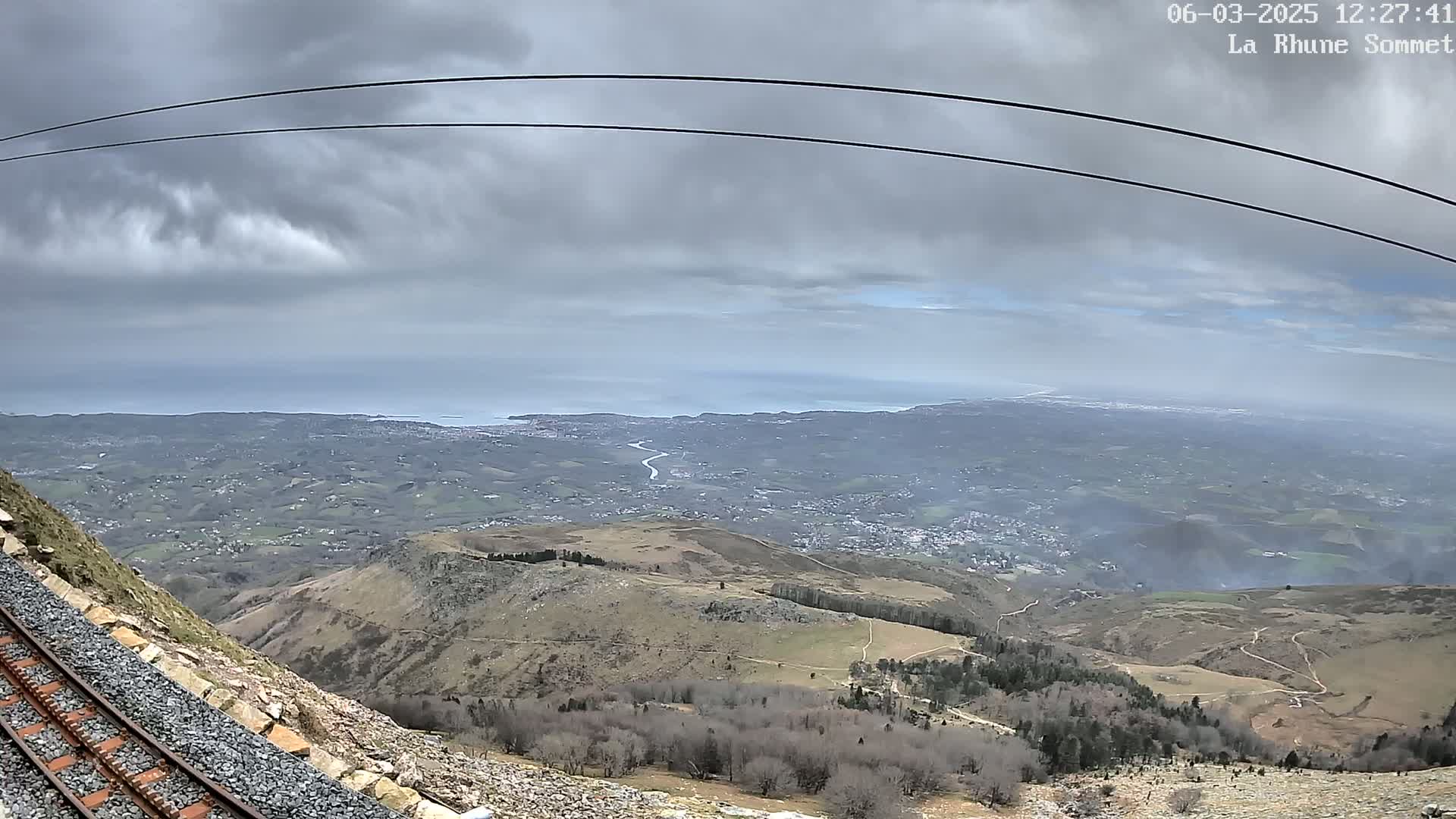 A mountaintop view under overcast skies shows a coastal landscape stretching to the sea, with a railway track running alongside a rocky hillside in the foreground.