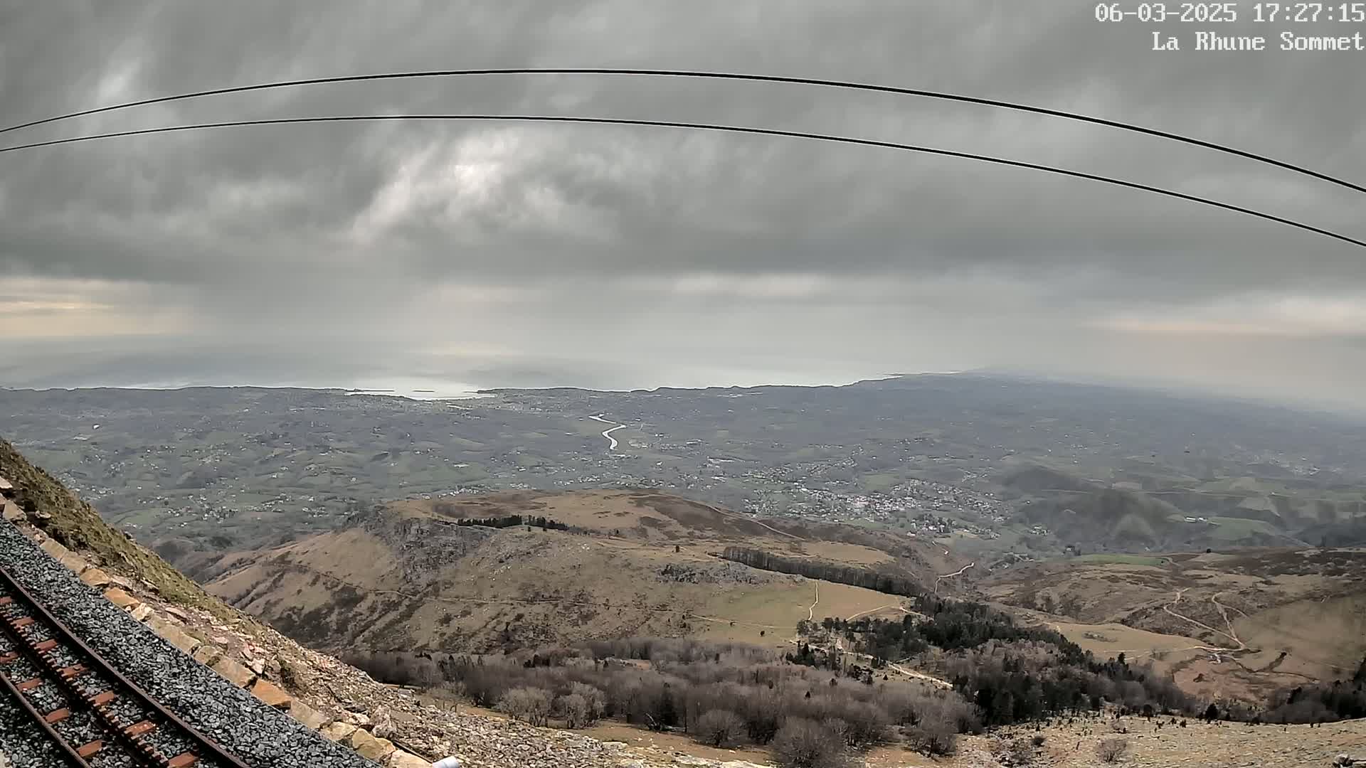 A mountaintop view reveals a railway track beside a rocky slope, overlooking a vast expanse of land leading to a grey, overcast sky and a distant ocean.