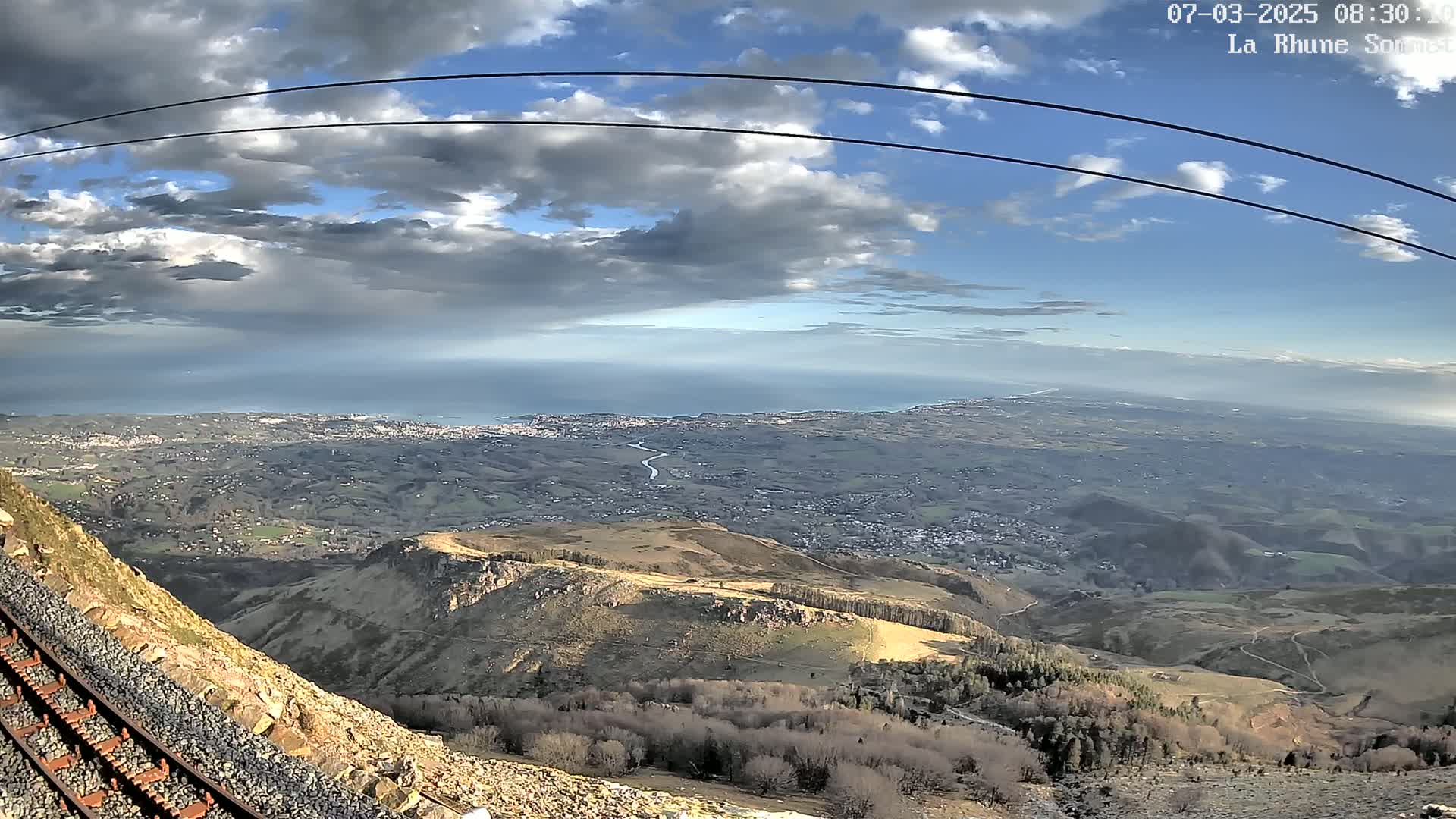A mountaintop railway track overlooks a sprawling coastal landscape under a partly cloudy sky.