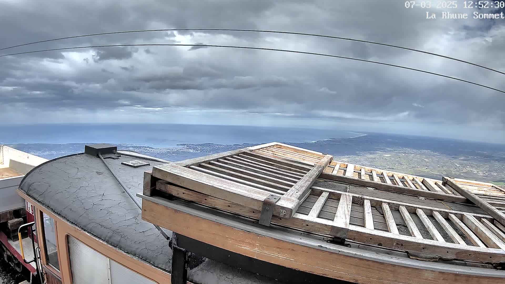 A portion of a train car with a weathered wooden gate on its roof overlooks a coastal landscape under a cloudy sky.