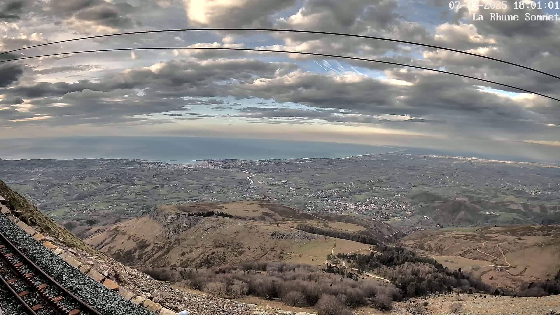 A mountaintop view shows a railway track beside a steep cliff overlooking a vast expanse of land, a distant ocean, and a cloudy sky.