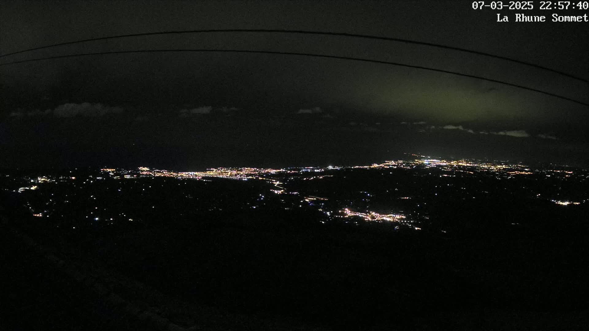 A nighttime, high-angle view shows a sprawling city illuminated by many small lights, partially obscured by darkness and a few clouds.