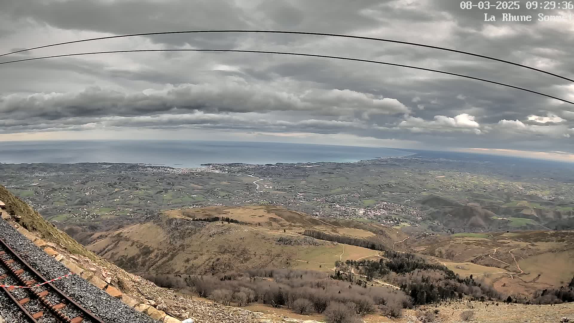 A mountaintop view under a cloudy sky shows a railway track beside a steep slope overlooking a vast expanse of land leading to a distant ocean.