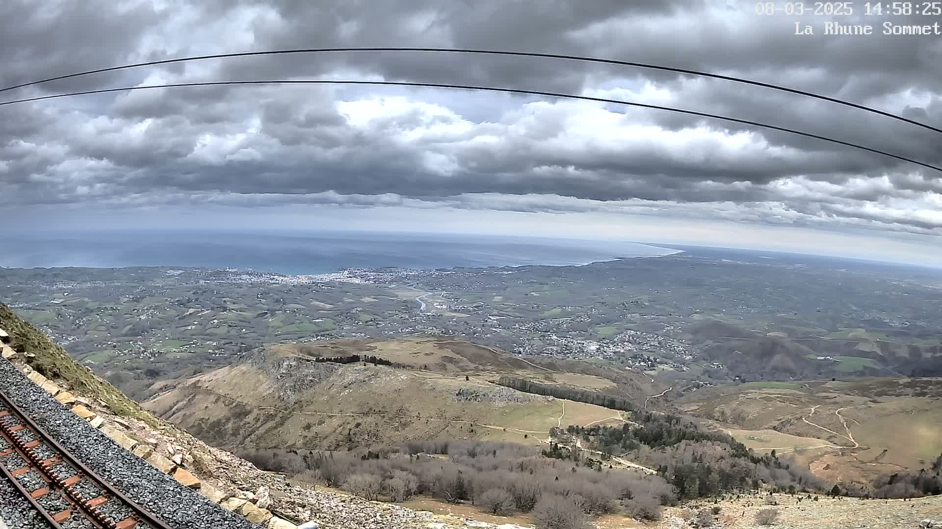 A mountaintop railway track overlooks a vast expanse of land, a distant ocean, and a cloudy sky.