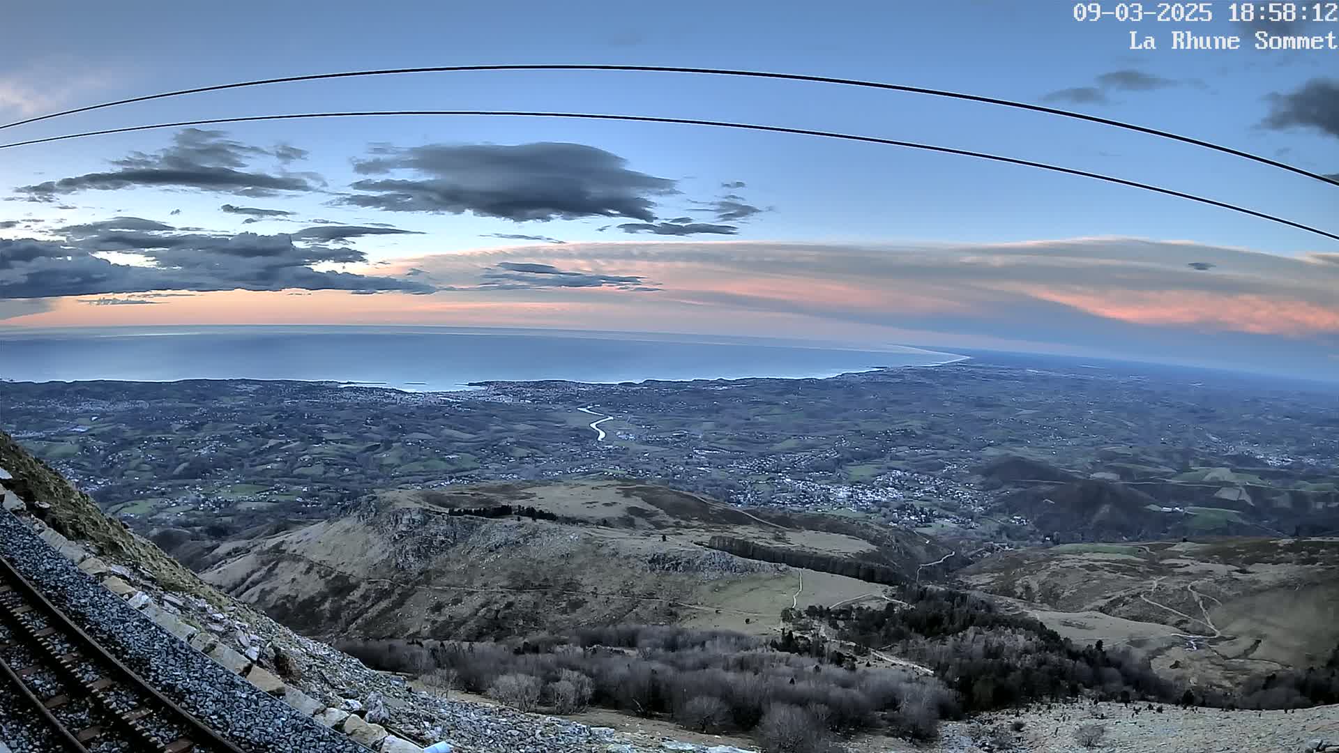 A mountaintop view at sunset shows a vast expanse of land and a calm sea in the distance, with a railway track alongside a rocky hillside in the foreground.