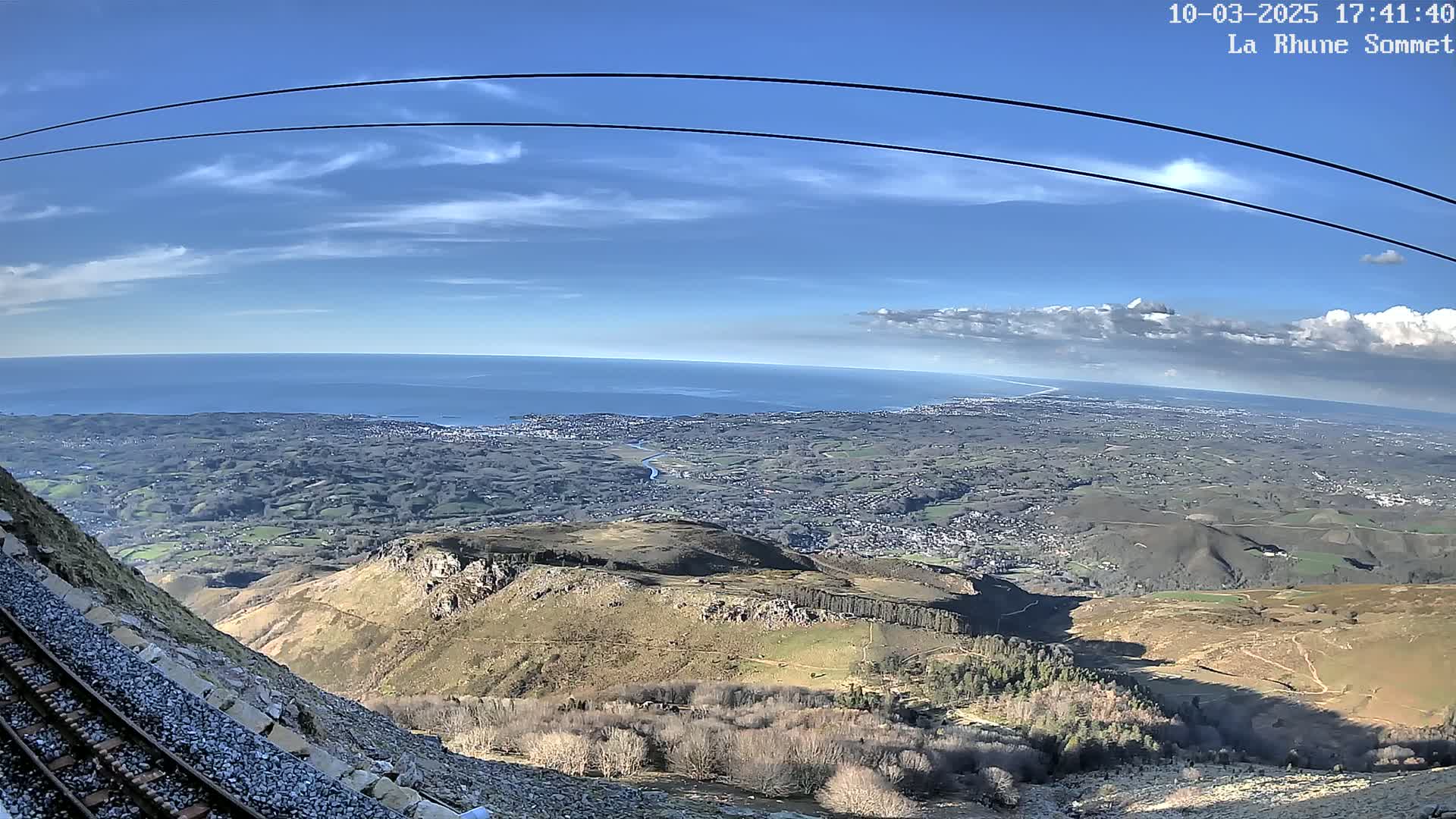A high-angle view from a mountaintop shows a vast expanse of land, a coastline with a calm ocean, and a railway track alongside a rocky slope under a mostly clear, blue sky with some clouds.