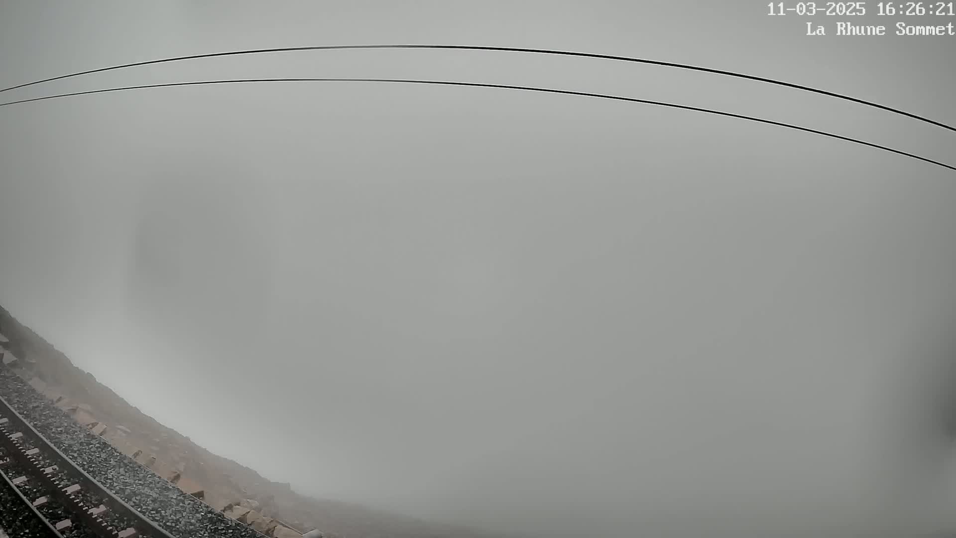 A low-visibility, foggy scene shows a train track next to a rocky embankment under an overcast sky.