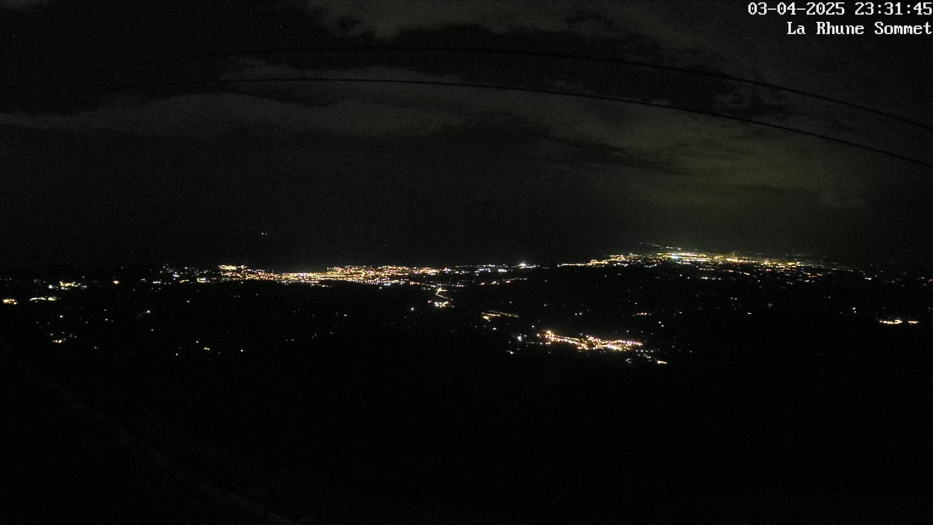 A nighttime, high-angle view shows a sprawling city's lights spread across a dark landscape under a cloudy sky.