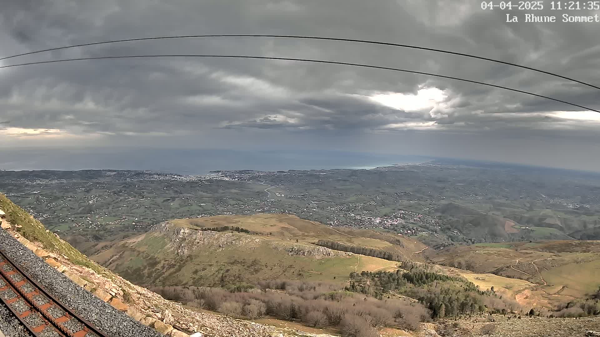 A mountaintop view under a cloudy sky shows a coastal town in the distance, with rolling hills and a railway track in the foreground.