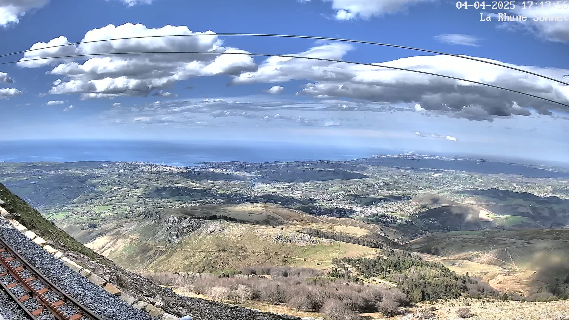 A mountaintop view shows a railway track alongside a steep hillside, overlooking a vast expanse of land leading to a distant ocean under a partly cloudy sky.