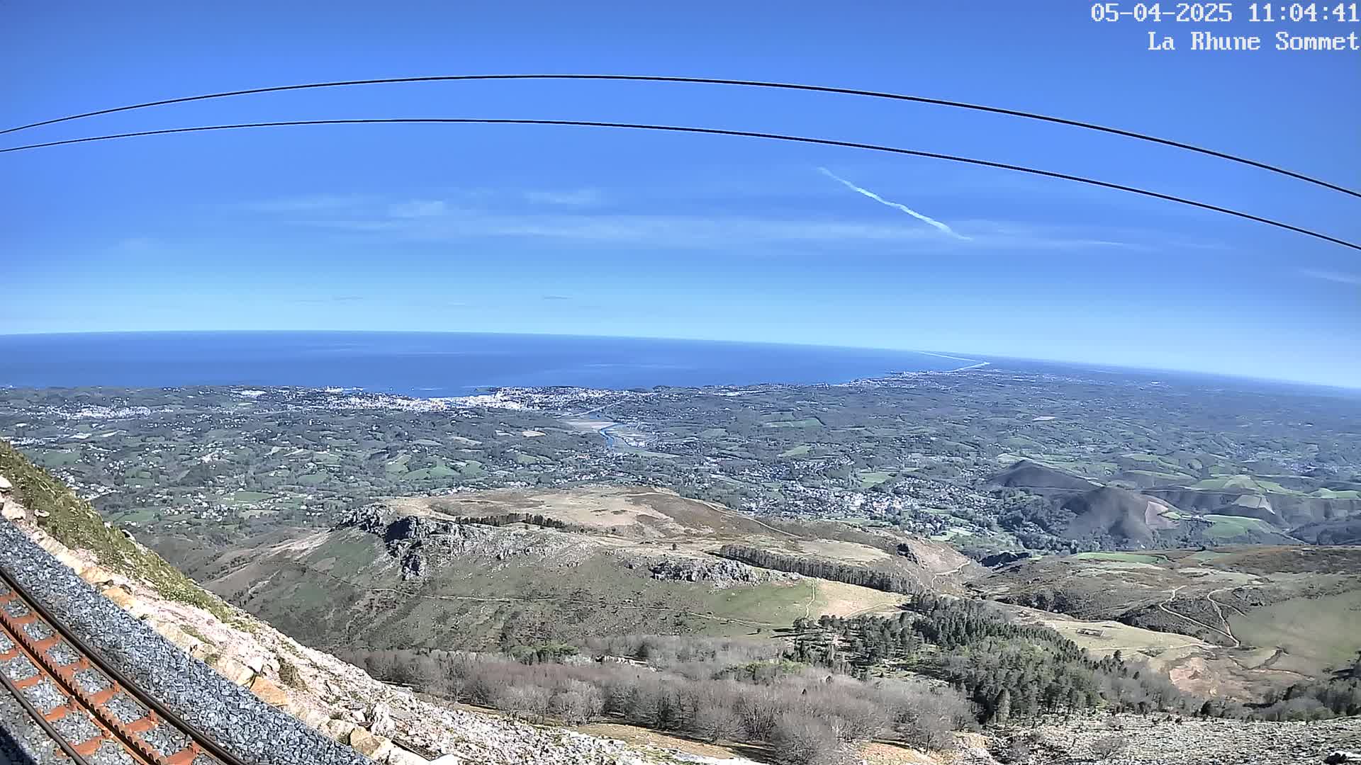 A mountaintop view on a clear, sunny day shows a vast expanse of ocean, coastal towns, and rolling hills, with a railway track visible in the foreground.