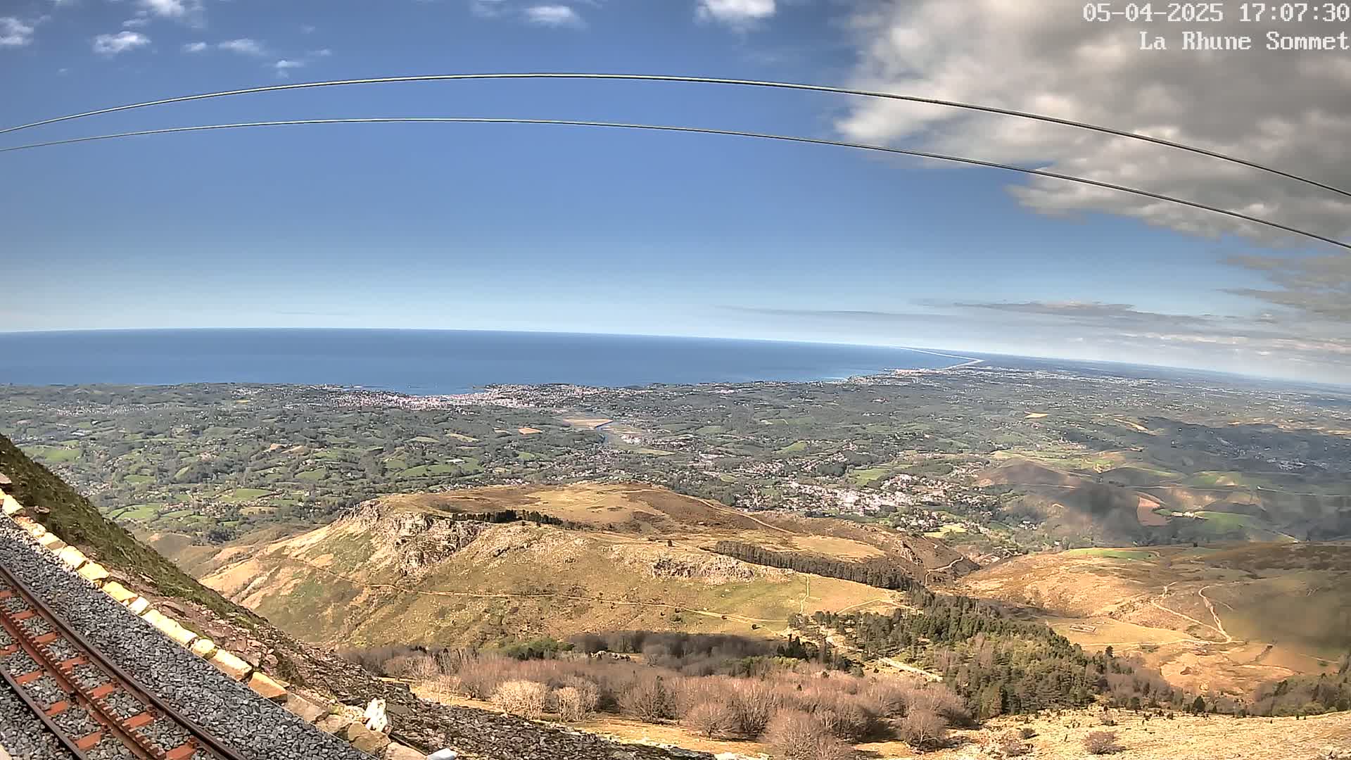 A mountaintop view on a partly sunny day shows a train track beside a rocky slope, overlooking a vast expanse of land leading to a blue ocean.
