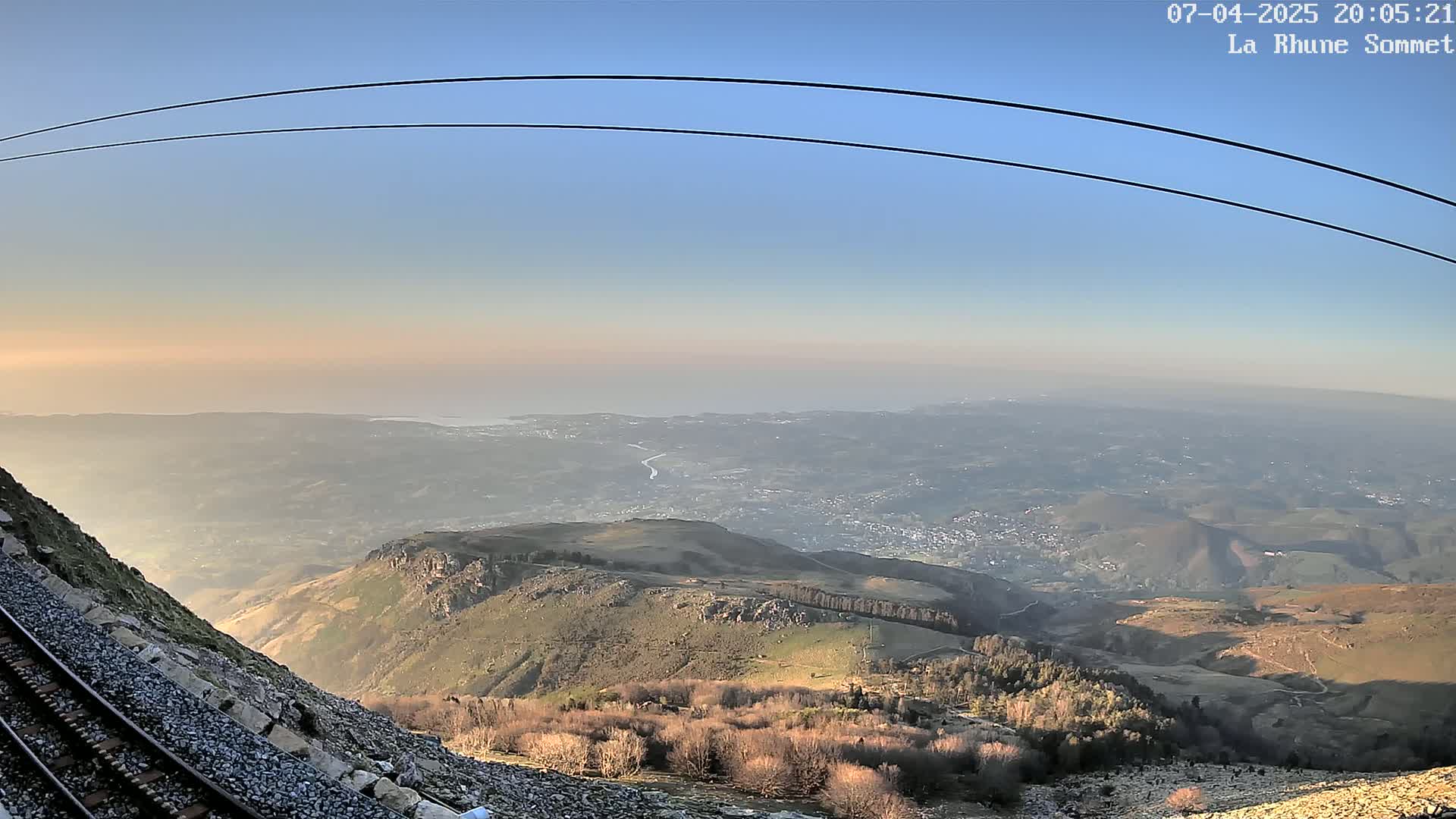A mountaintop view at sunset shows a train track beside a rocky slope, overlooking a vast expanse of land stretching to a hazy coastline under a clear sky.