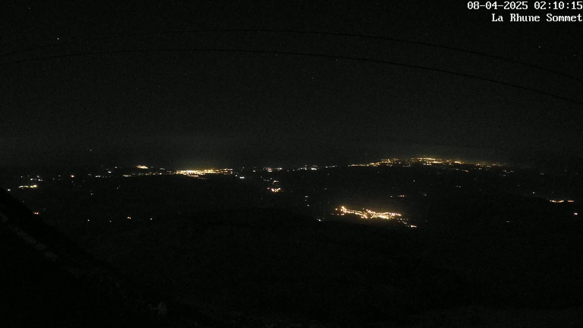 A nighttime, high-angle view shows distant city lights spread across dark, mountainous terrain under a clear, star-filled sky.